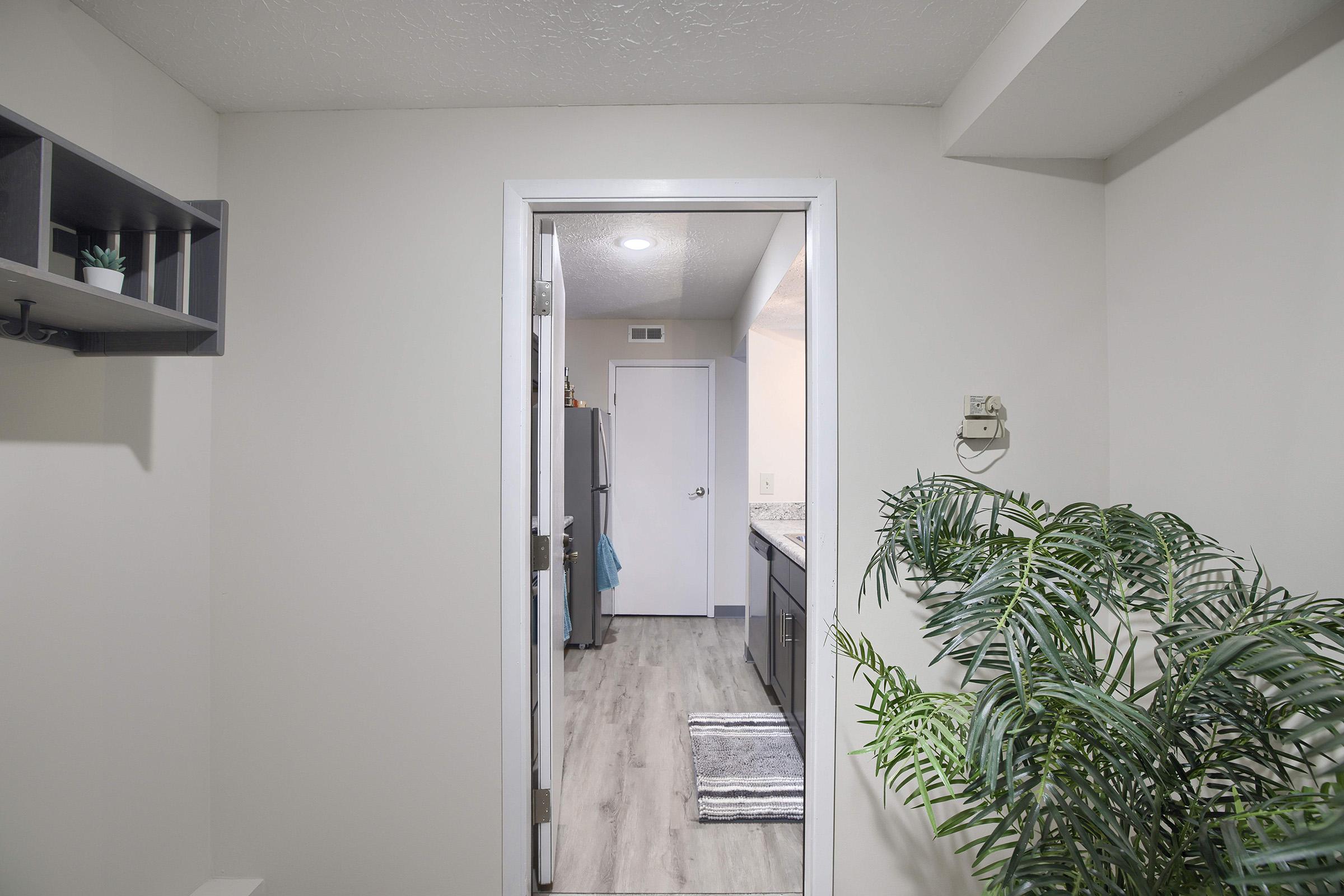 A view through an open door into a kitchen, featuring light-colored walls, gray cabinetry, and modern appliances. A potted plant is visible on the right side, and the floor has a wood-like finish. The space appears clean and well-lit with overhead lighting.