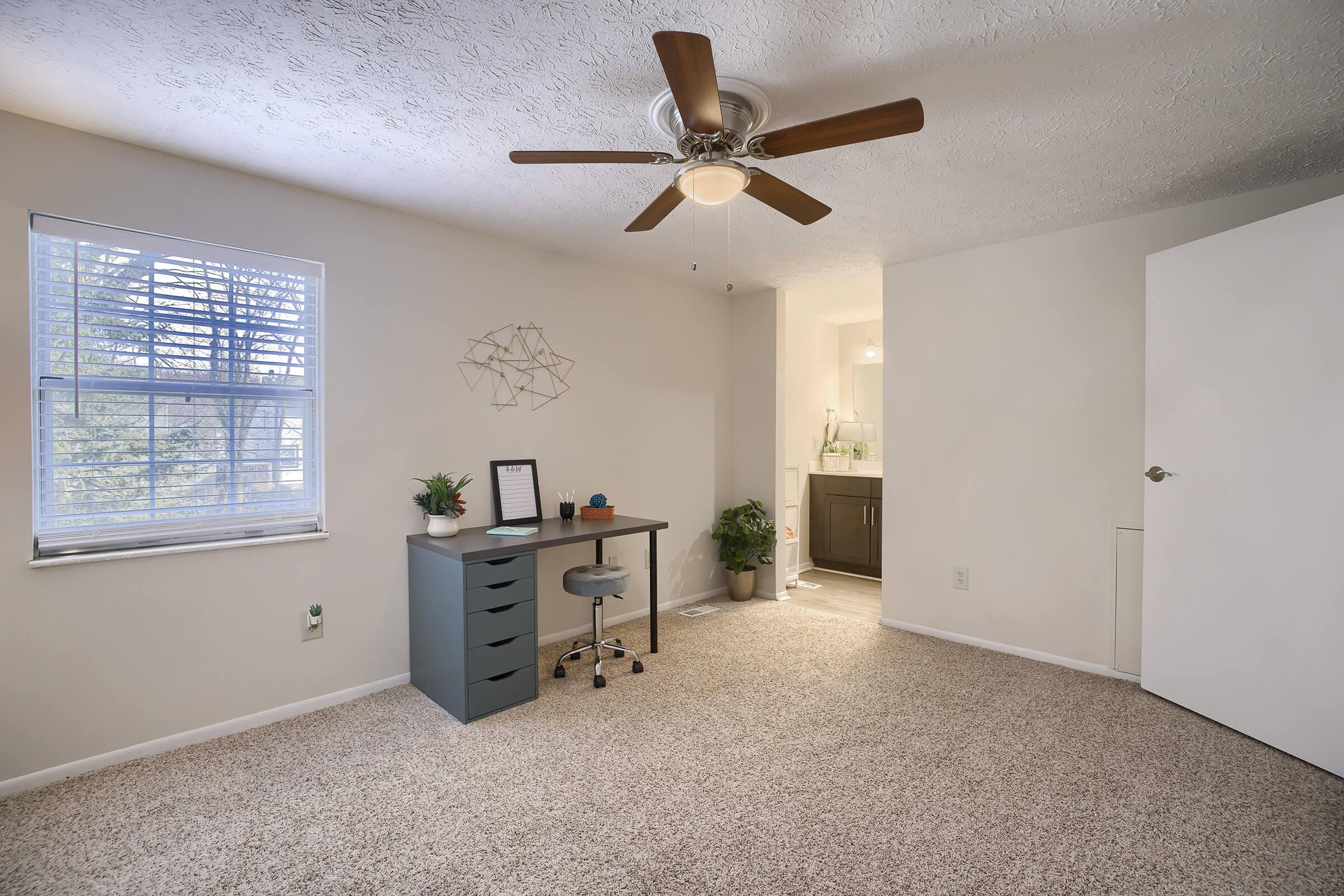 A contemporary room with beige carpet and light gray walls, featuring a window with blinds letting in natural light. There is a small desk with a chair and decorative plant, and a closet to the right. A modern ceiling fan is overhead, and a bathroom area is visible in the background.