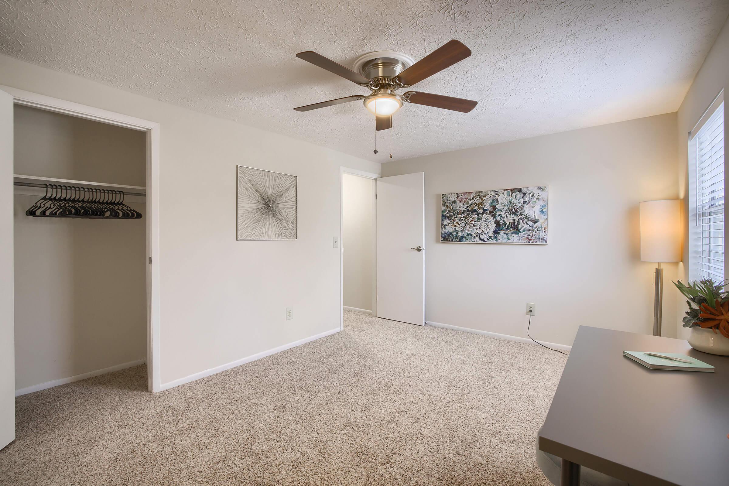 A neatly arranged bedroom featuring light-colored walls and beige carpet. There's a ceiling fan with five blades, a closet with hanging space, an abstract artwork on the wall, a decorative light lamp, and a small desk with a plant. Natural light streams in through a window with blinds.