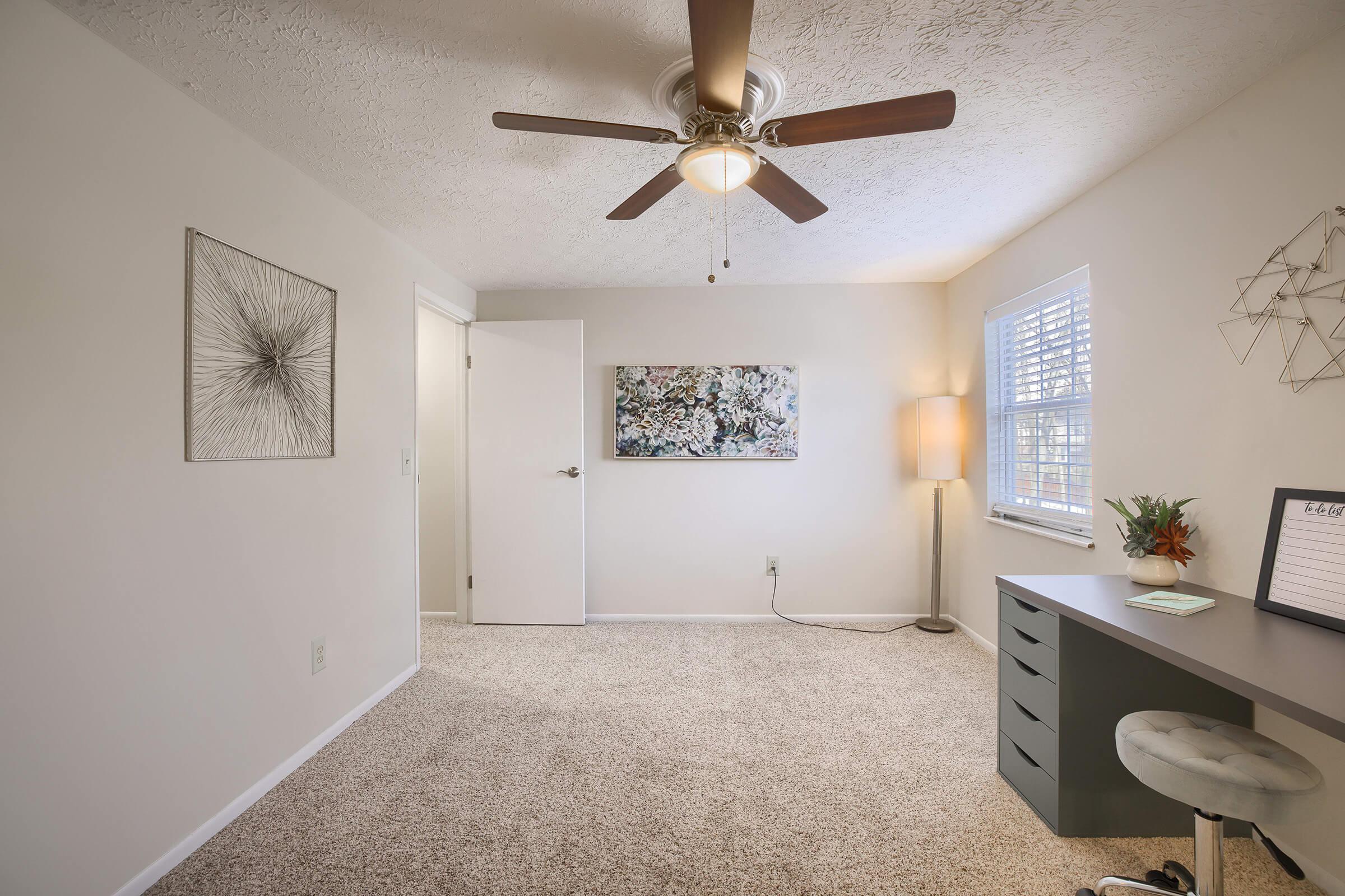 A well-lit, minimalist room featuring a ceiling fan, light-colored walls, and carpeted flooring. There is a small desk with storage, a modern lamp, and a decorative wall art piece. A large window allows natural light to enter, enhancing the airy feel of the space.