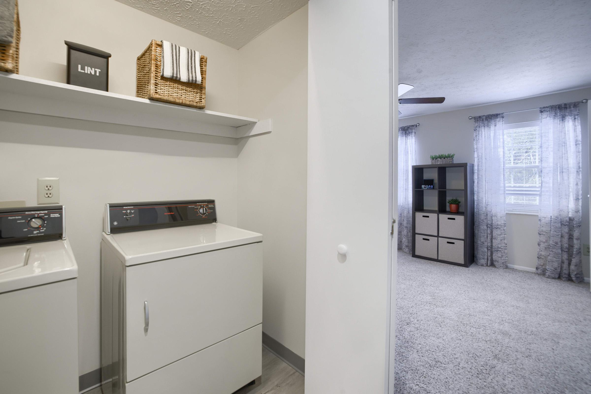 A laundry room featuring a white washing machine and dryer with a shelf above displaying a lint container and baskets. In the background, a light and airy living space with a window covered by sheer curtains and a shelf unit filled with cubed storage boxes.