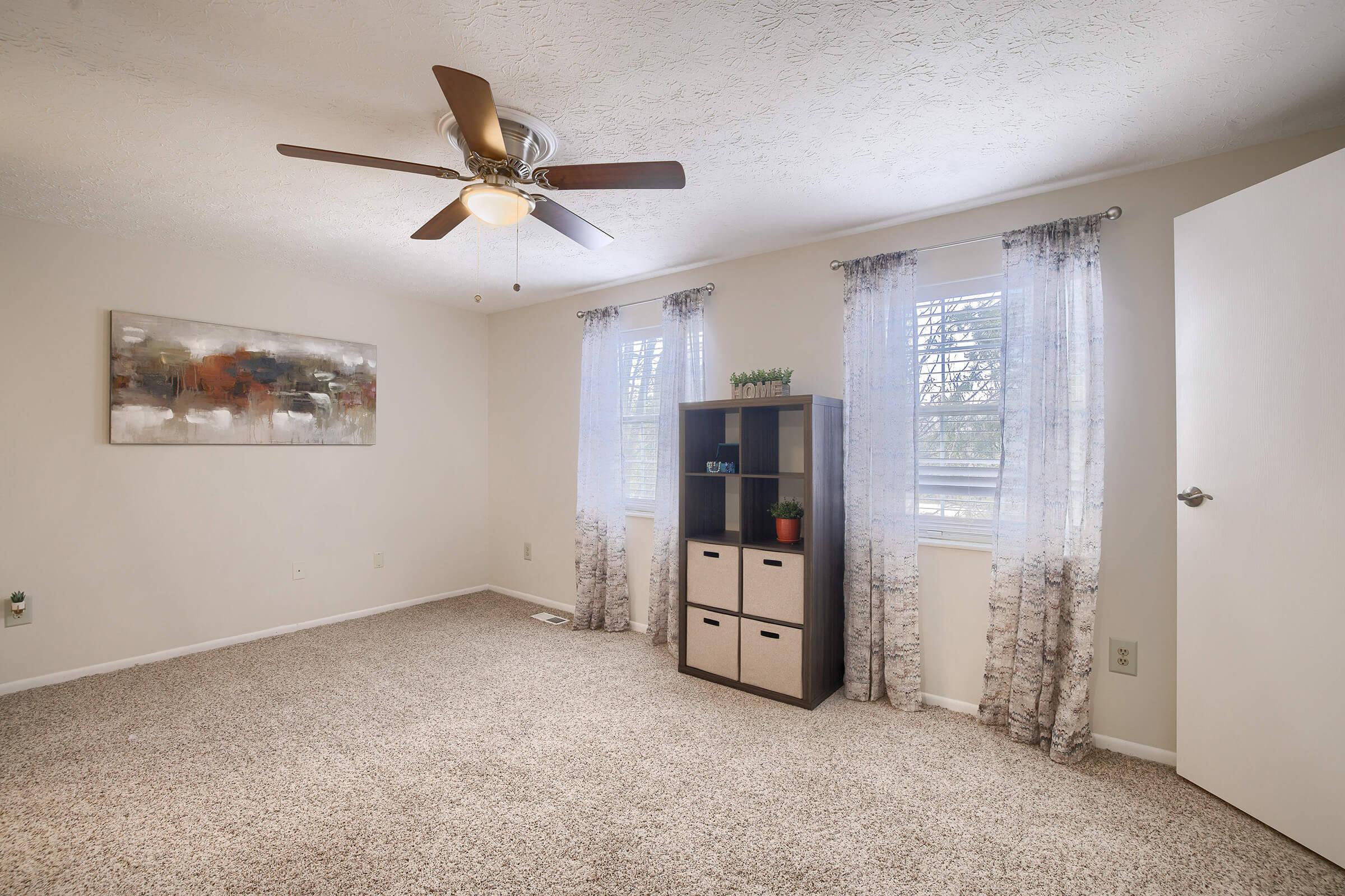 A well-lit room featuring beige walls and light-colored carpet. There's a ceiling fan with wooden blades, large windows with sheer curtains, and a small black and beige storage unit against the wall. An abstract painting hangs above the unit, adding a touch of decor to the space.