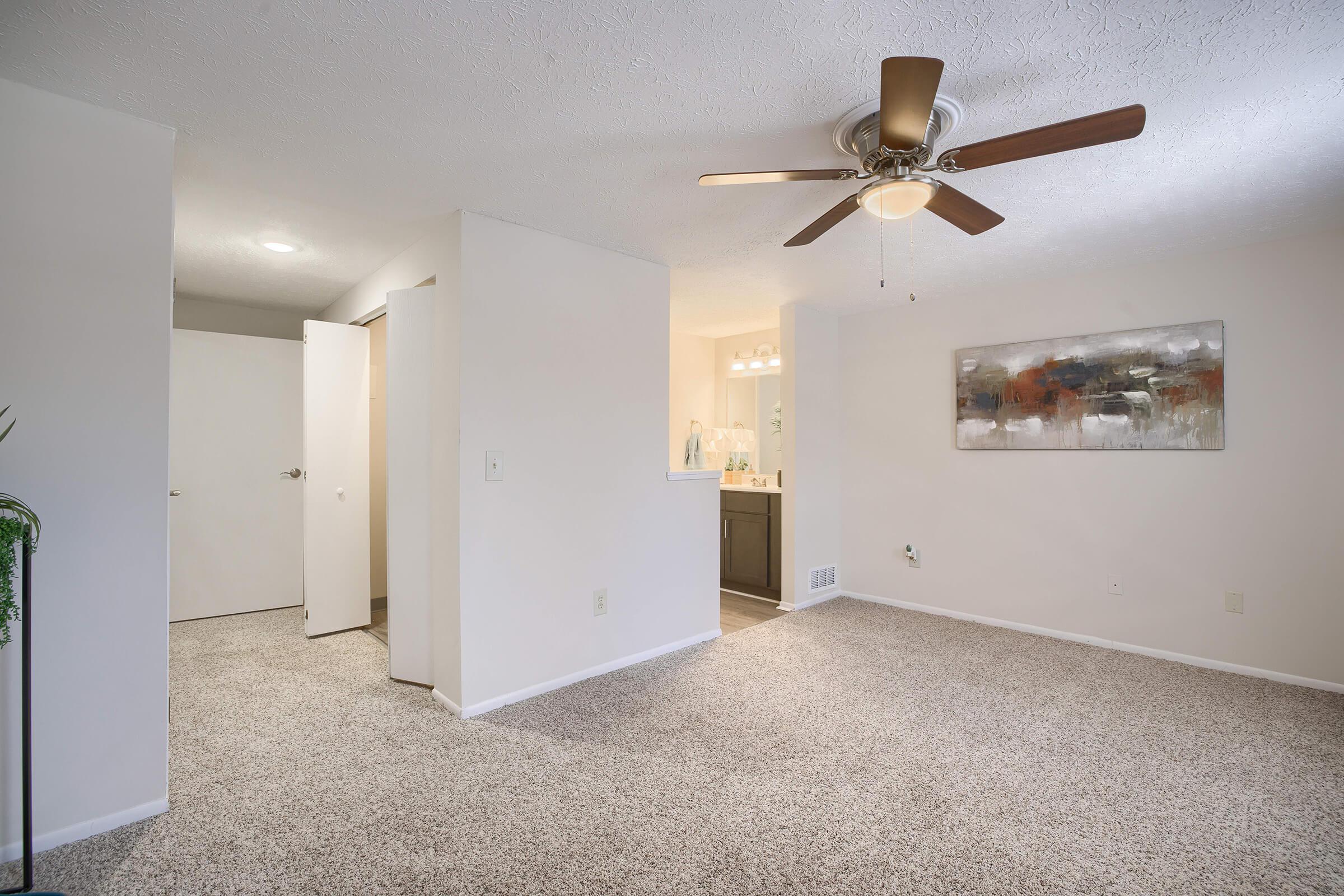A spacious, well-lit living area featuring light beige walls and a textured carpet. A ceiling fan with wooden blades hangs above. To the left, an open door leads to another room. The background includes a glimpse of a kitchen area with modern fixtures, and a decorative painting hangs on the right wall.