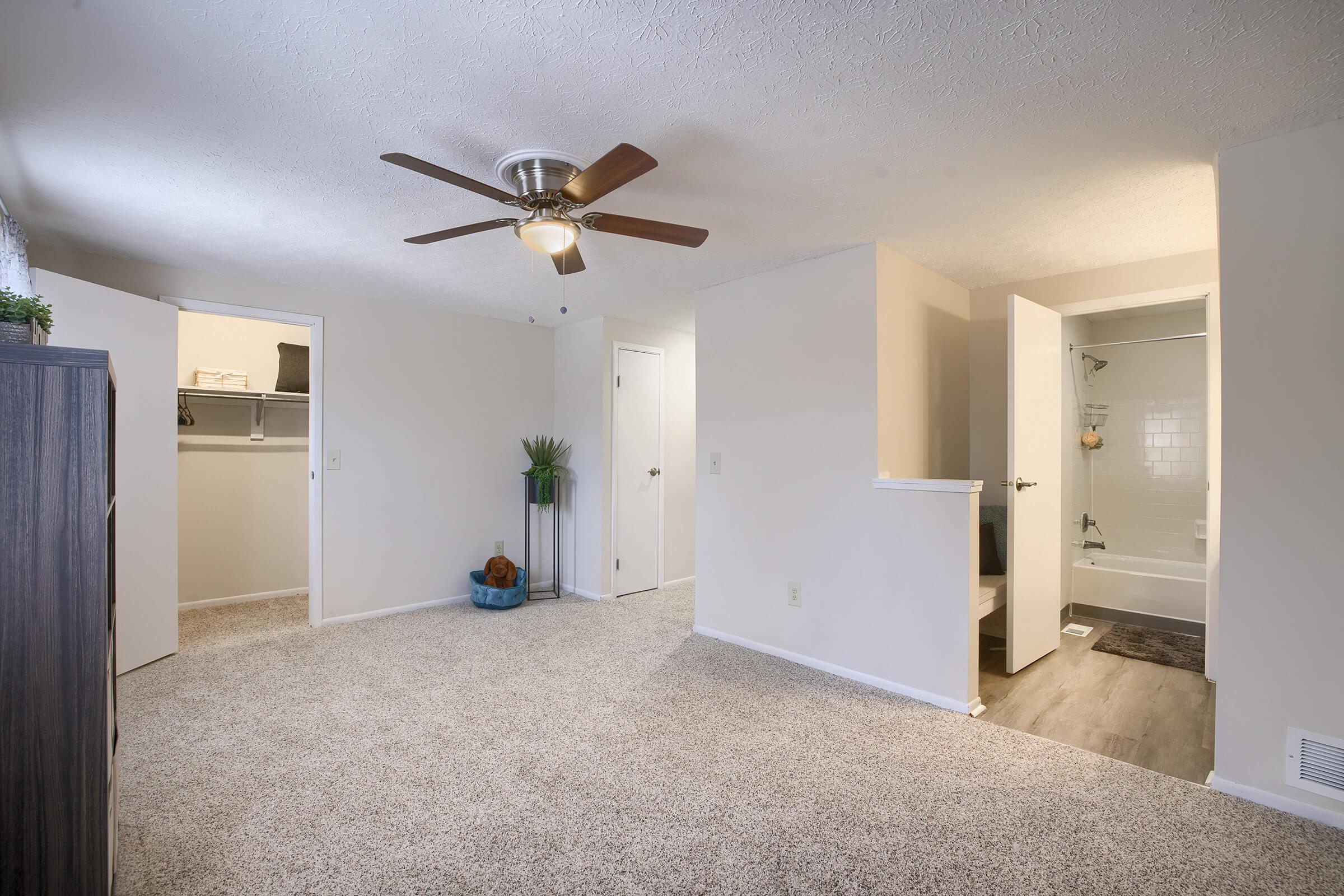A spacious, well-lit living area with light-colored walls and carpet. It features a ceiling fan and an open layout, leading to a small bathroom visible in the background. A closet is on the left, and decorative plants add a touch of greenery to the space.