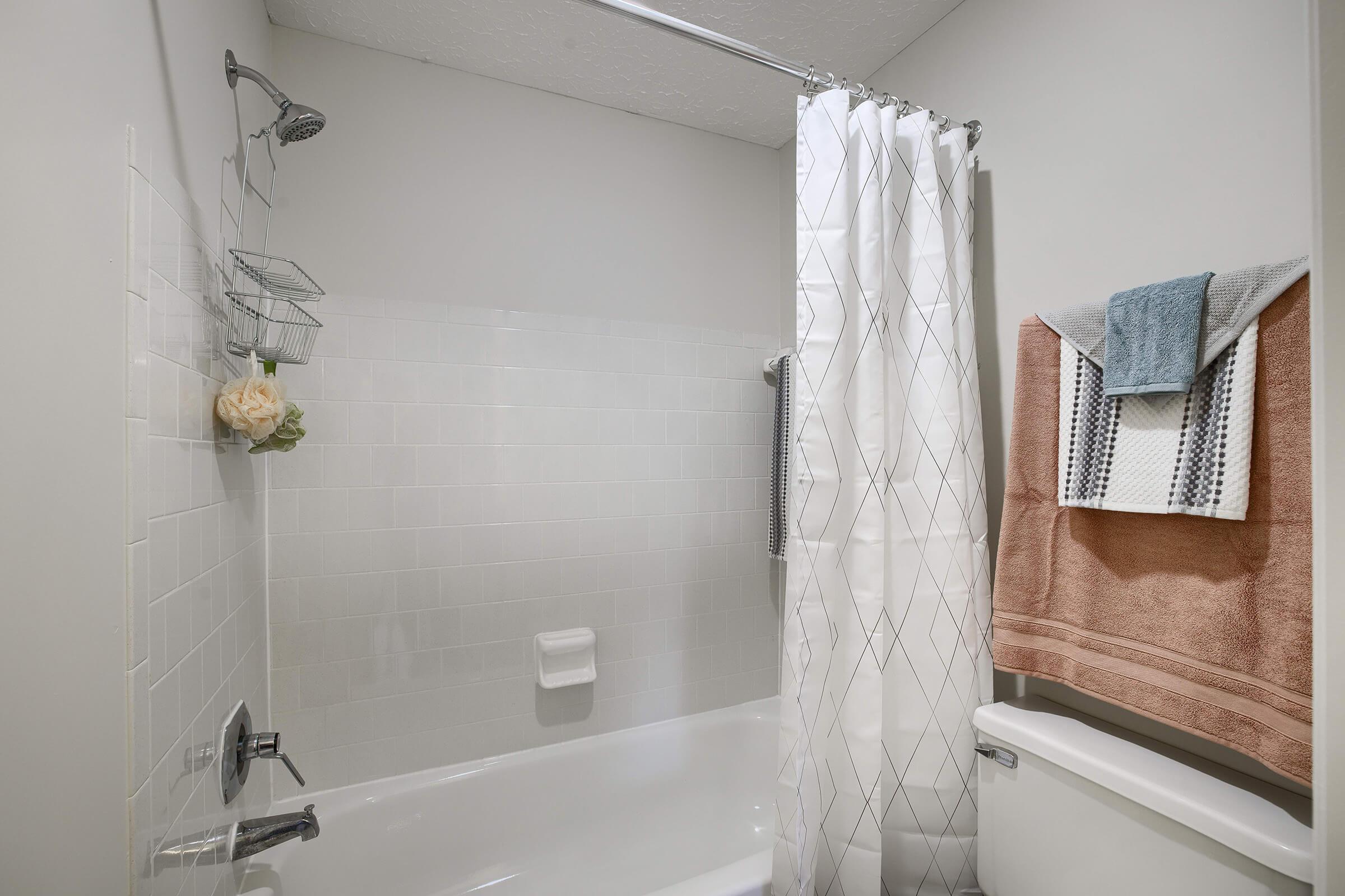 A clean bathroom with a shower area featuring a white curtain with a geometric pattern. There are wire shelves holding toiletries, a light-colored towel hanging on a bar, and a light brown towel folded on top of the toilet tank. The walls are light-colored tiles, and the space is bright and well-maintained.