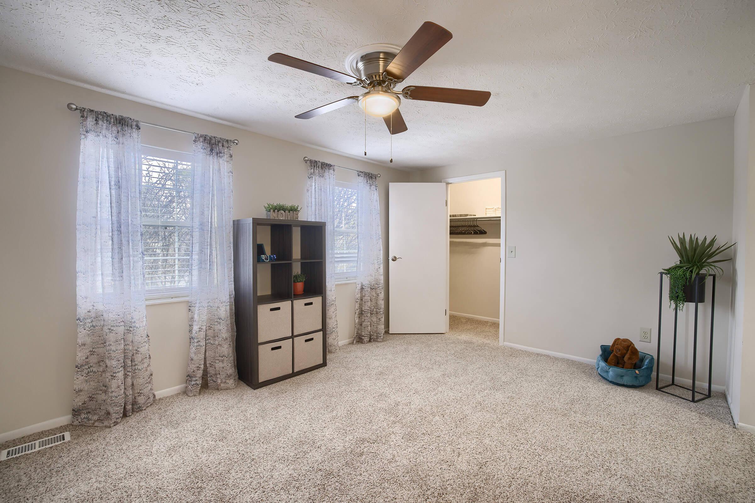 A bright, spacious room featuring light-colored walls, a ceiling fan, and large windows with sheer curtains. There's a bookshelf with storage cubes, a small potted plant, and a decorative item in a blue container. The carpeted floor adds warmth to the space, creating a cozy atmosphere.