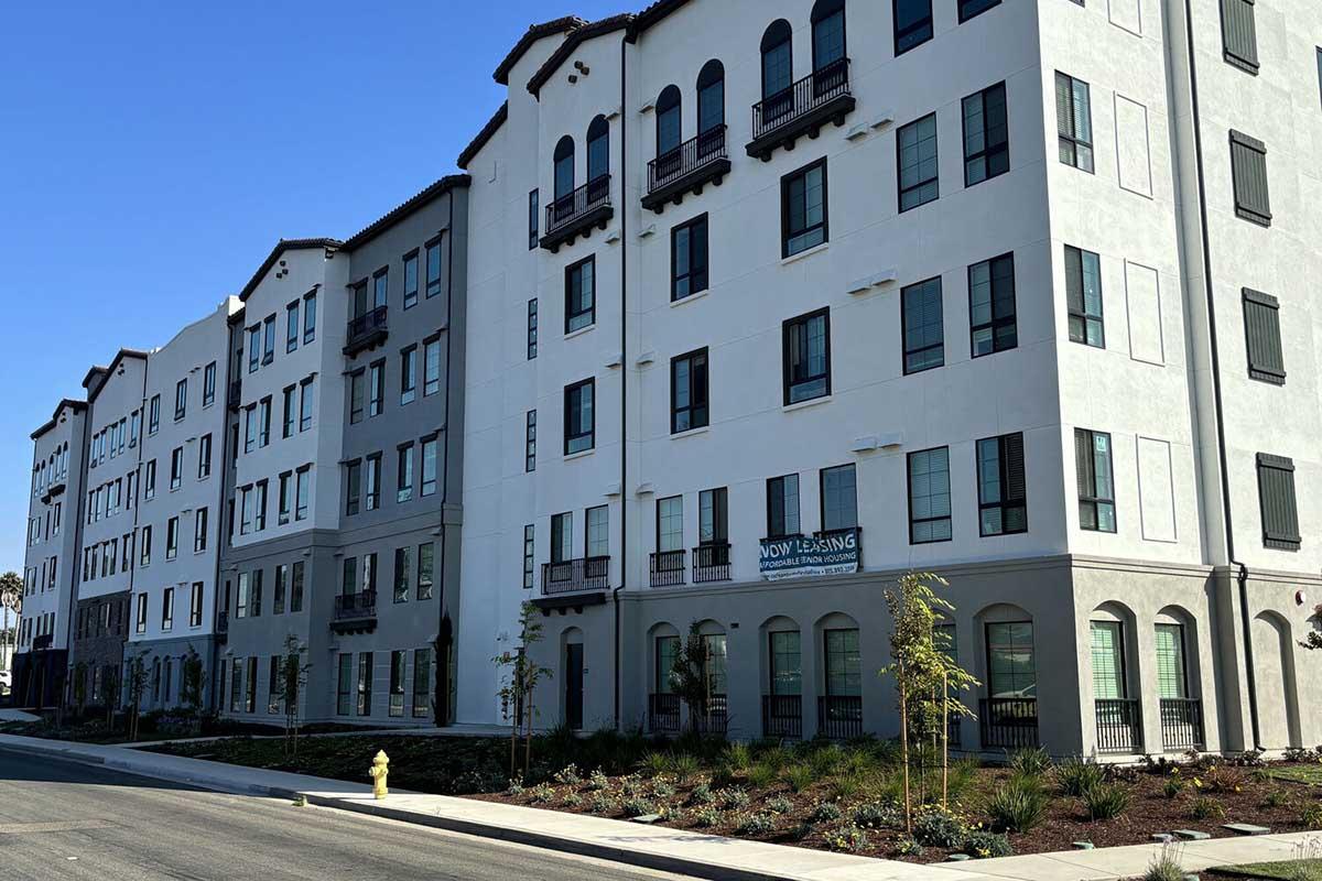 A modern multi-story residential building with a clean white exterior, featuring multiple windows and balconies. The foreground shows a landscaped area with small trees and shrubs, along with a fire hydrant on the left side of the image. A sign indicates that the building is available for leasing.