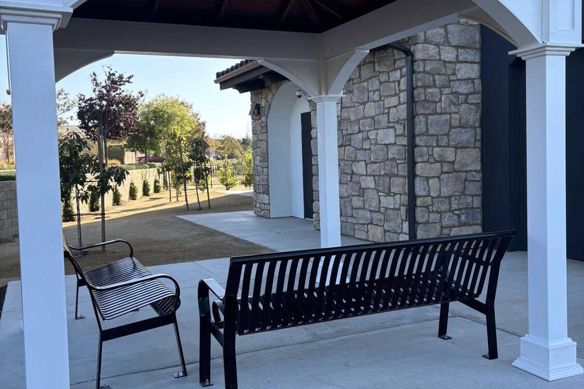 A black metal bench sits beneath a white gazebo-like structure, flanked by stone walls. In the background, trees are visible along a pathway, with well-maintained grass, creating a serene outdoor setting. The scene is bright and inviting, suggesting a peaceful place to sit and relax.
