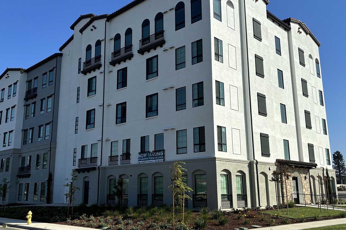 Newly constructed multi-story residential building with a modern design. The exterior features a mix of white stucco and dark accents. Large windows are prominent, and there is a "Now Leasing" sign displayed. Surrounding landscaping includes small trees and shrubs, and the sky is clear and blue.