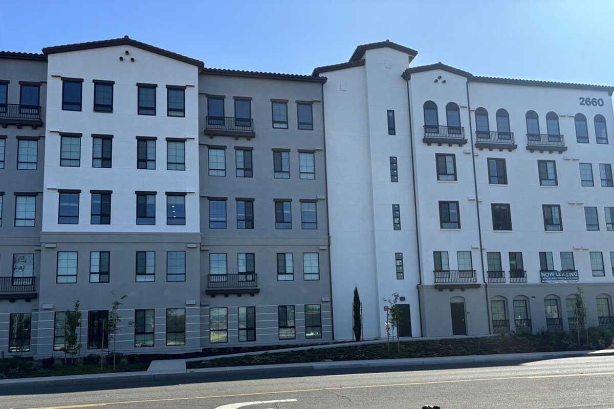 A modern multi-story building with a mix of gray and white exteriors, featuring large windows and balconies. The structure has a distinctive architectural style with decorative elements. It's located near a road, with some greenery in front.