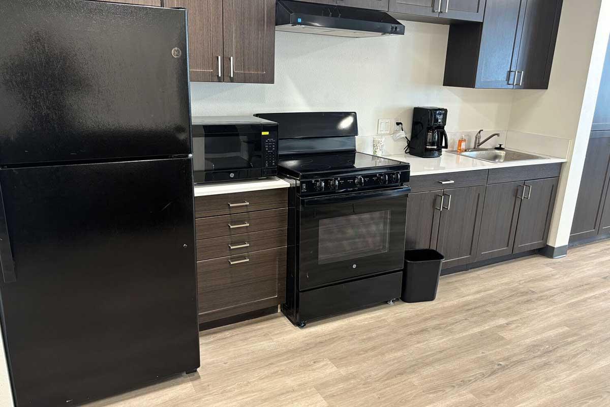 A modern kitchen featuring a black refrigerator, a black microwave, a black gas stove with an oven, and a stainless steel sink. A coffee maker is on the counter next to the stove, and a small trash bin is located beside the stove. The cabinetry is dark wood, and the flooring is light laminate.