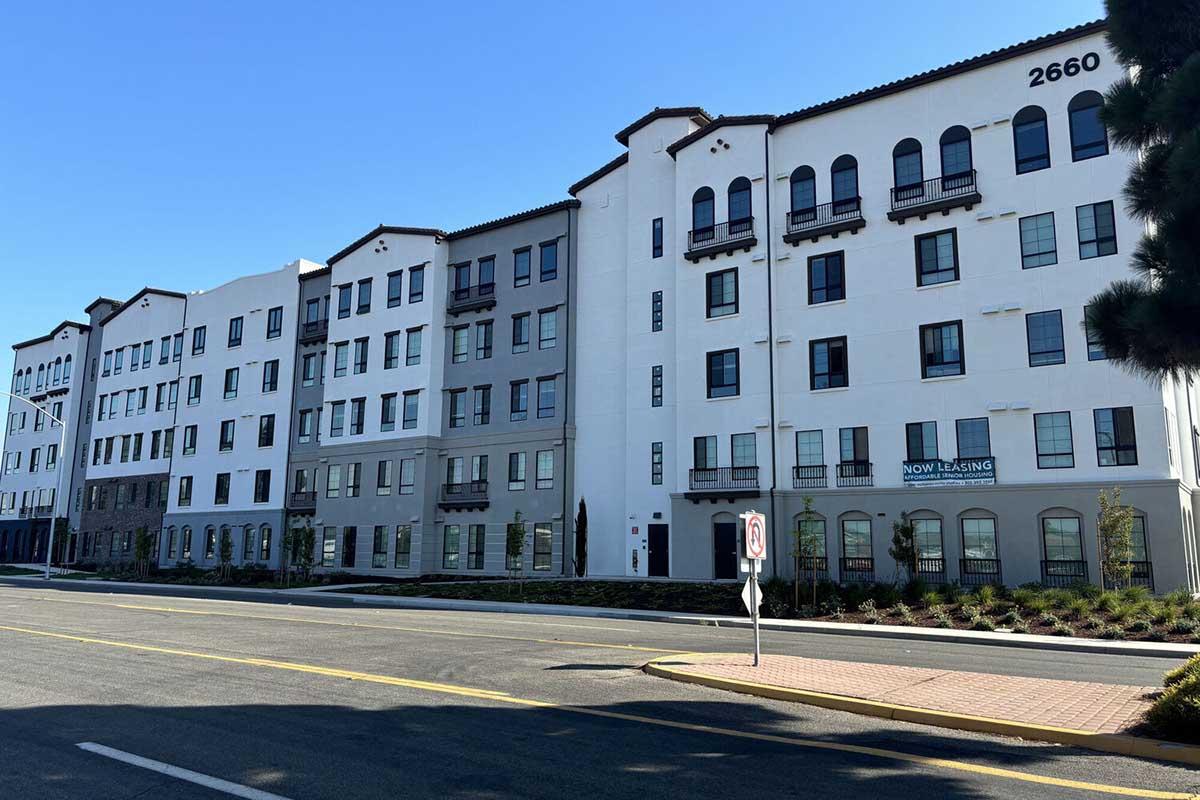 A modern multi-story apartment building featuring a mix of white and gray exteriors. The building has numerous windows and balconies, with a sign indicating "NOW LEASING." A landscaped area and street can be seen in front, along with a no parking sign. The setting is sunny and clear.