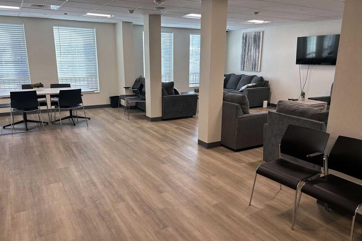 A spacious, modern lounge area featuring a seating arrangement with gray sofas, a coffee table, and several chairs. A dining table with chairs is on one side, while a flat-screen TV is mounted on the wall. There are large windows allowing natural light to fill the room, and the floor is covered with light-colored wood laminate.
