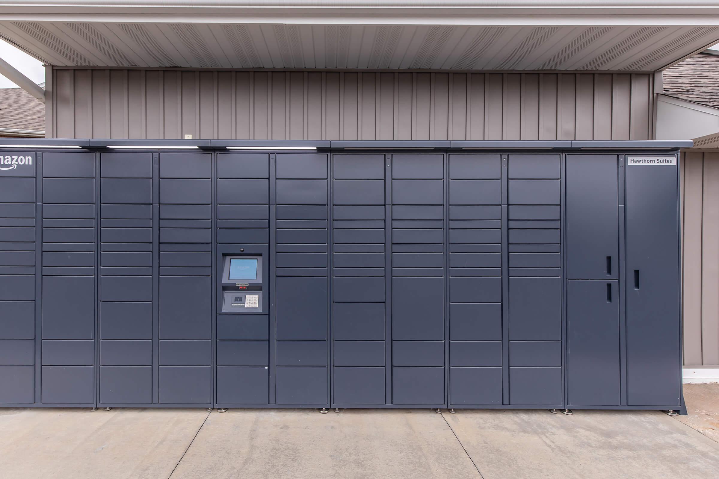 A row of Amazon delivery lockers placed against a beige wall, with an electronic touch screen on one of the lockers for package retrieval. The design is modern and sleek, suitable for outdoor use in a residential or commercial area.