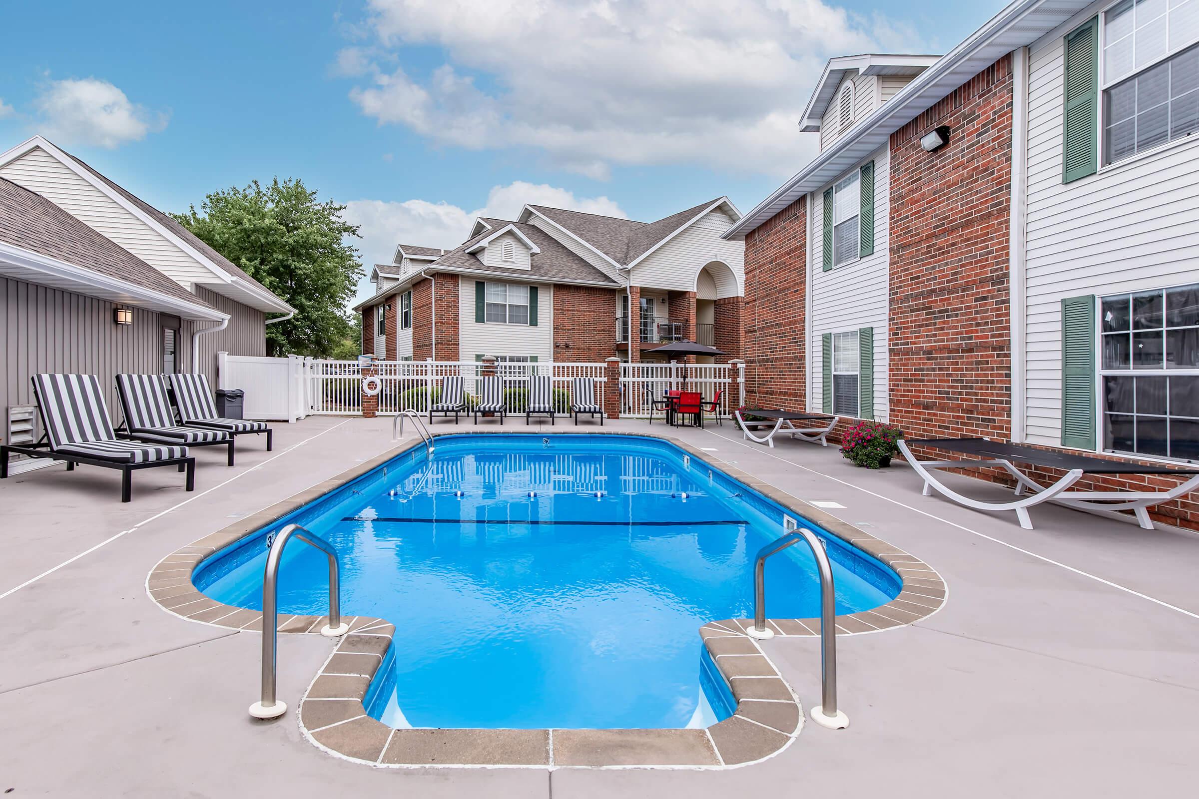 A clear swimming pool surrounded by lounge chairs and hammocks, set in a sunny outdoor area. The pool is located near brick-clad buildings with white accents. Lush greenery is visible in the background, creating a relaxing atmosphere.