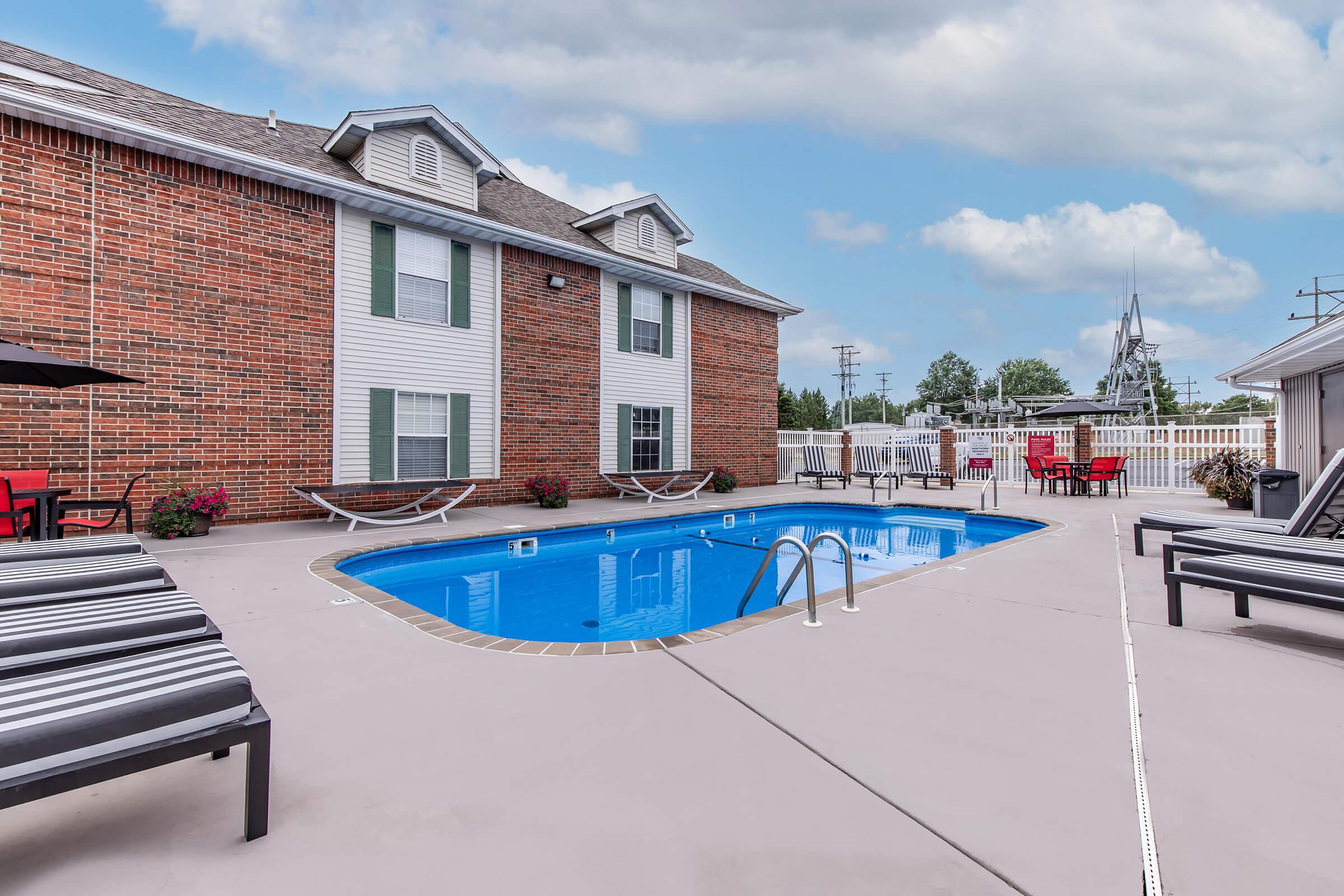 A residential swimming pool area featuring a blue pool with a ladder, surrounded by lounge chairs, umbrellas, and potted plants. The backdrop includes a brick building with windows and a white fence, set against a partly cloudy sky.