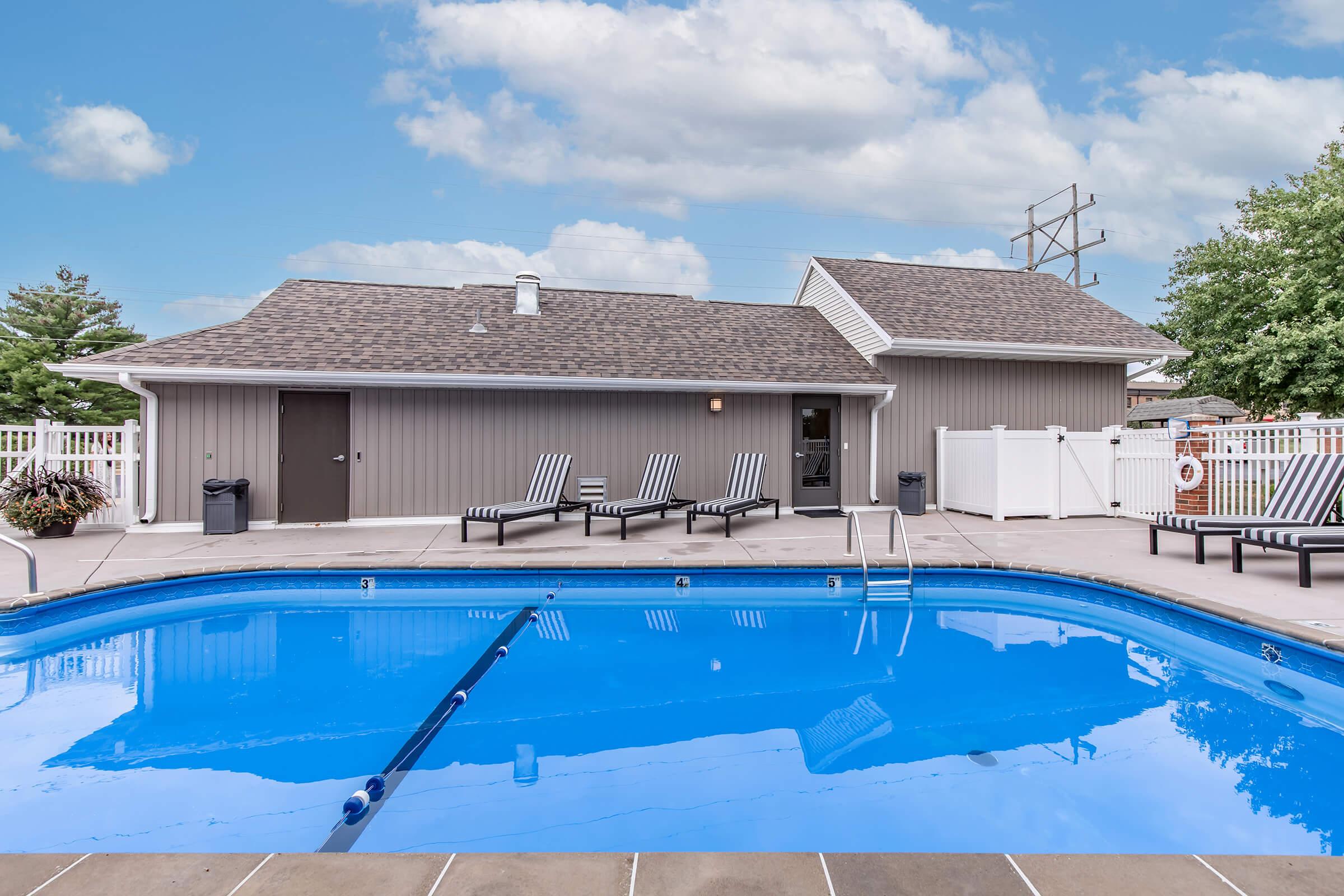 A clear blue swimming pool surrounded by lounge chairs under a bright sky. In the background, there is a single-story building with a gray exterior and a fenced area. The pool area is well-maintained with decorative plants and safety equipment nearby.