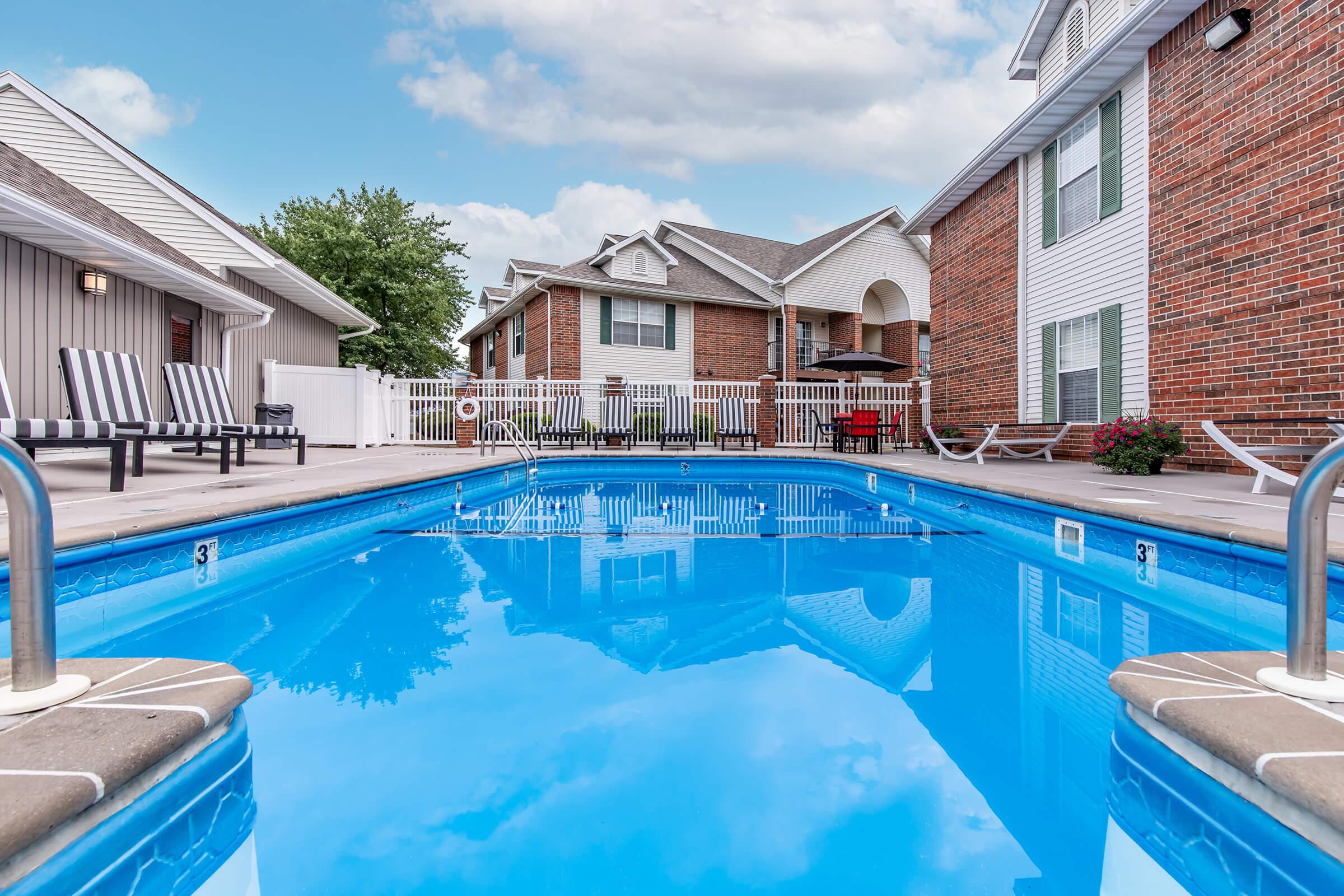 A clear blue swimming pool reflects the sky, surrounded by a beige deck. In the background, red-brick buildings with white trim are visible, along with patio furniture and landscaping. The scene is serene, with a few poolside loungers and a shaded seating area under an umbrella.