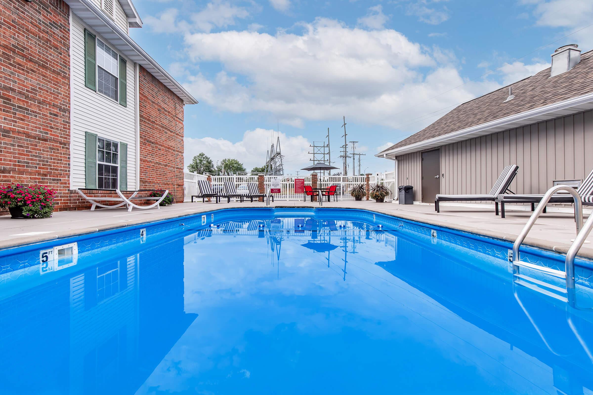 A clear blue swimming pool surrounded by a patio, with lounge chairs and colorful flowers along the edge. In the background, there is a building and a view of sailboats moored nearby under a partly cloudy sky.