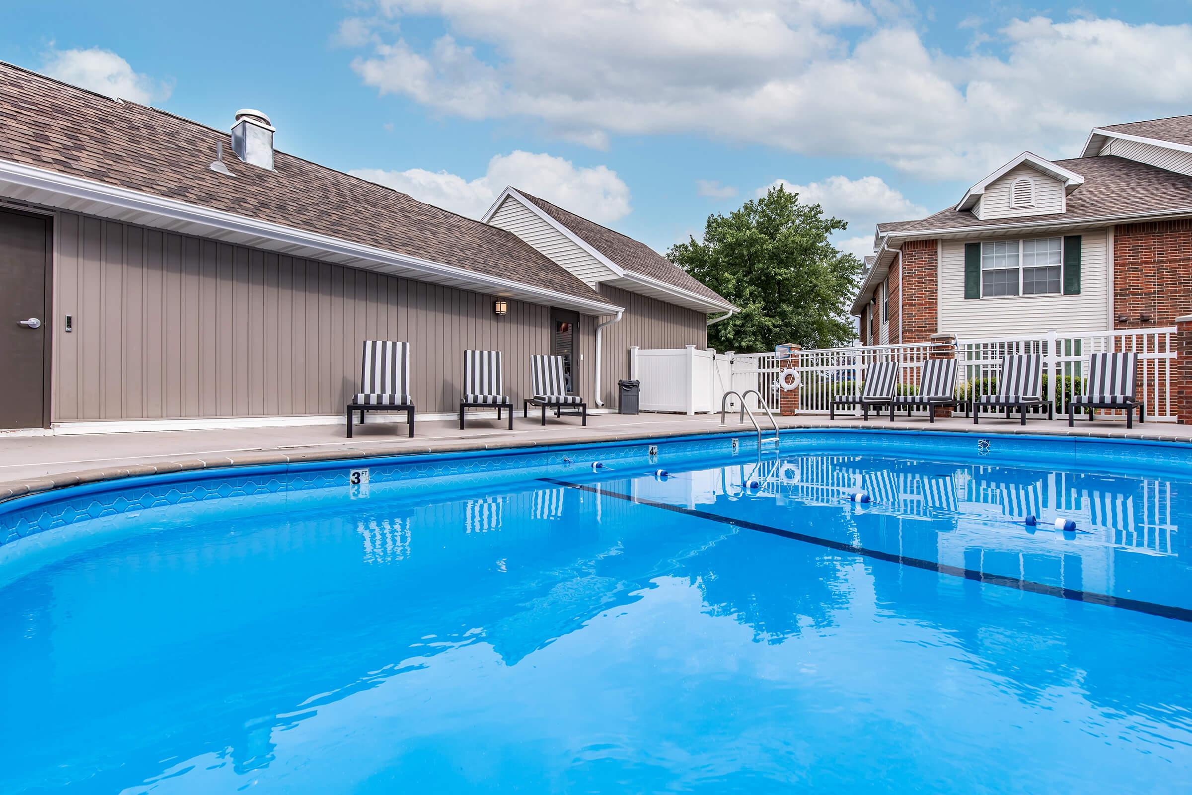 A clear blue swimming pool surrounded by a patio area with lounge chairs. In the background, there are two residential buildings with a fence, trees, and a partly cloudy sky above. The pool area looks inviting and well-maintained, perfect for relaxation and leisure activities.