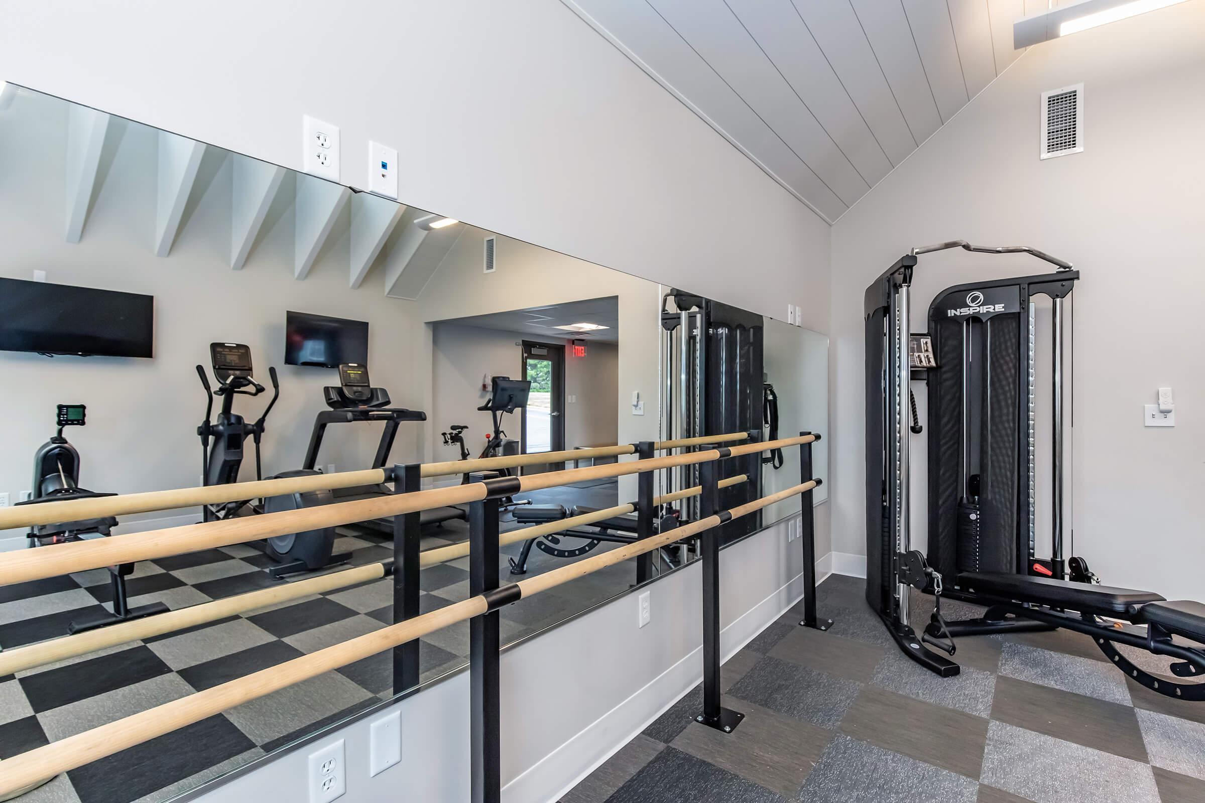 A modern fitness studio featuring gym equipment like treadmills and ellipticals, along with a mirrored wall and a cable machine. There is a barre installed along one wall, and the space has a clean design with gray flooring and ample natural light.
