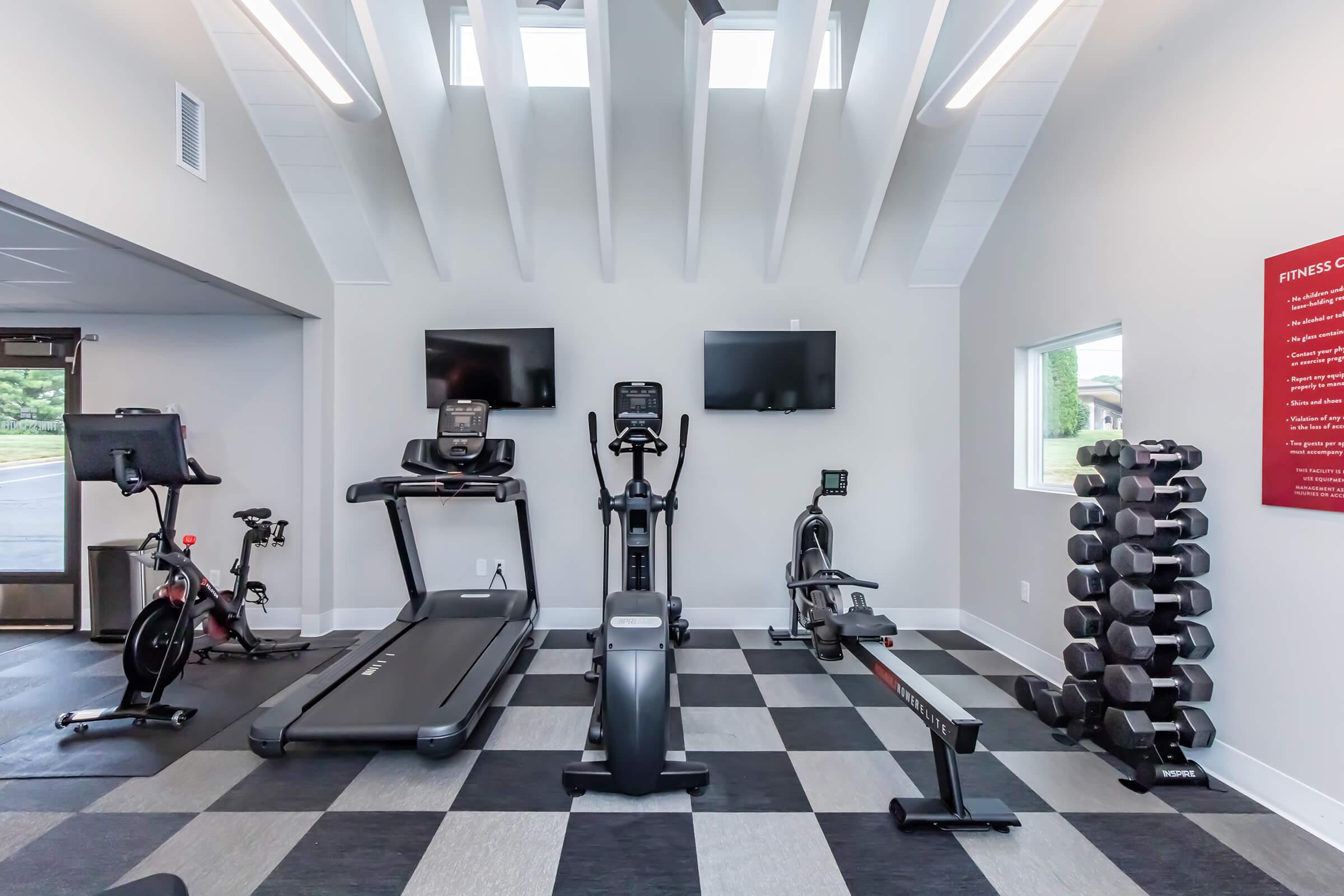 A modern fitness room featuring various exercise equipment, including a treadmill, elliptical machine, stationary bike, rowing machine, and free weights. The space has large windows for natural light and two flat-screen TVs mounted on the wall. The flooring is a combination of light and dark tiles.