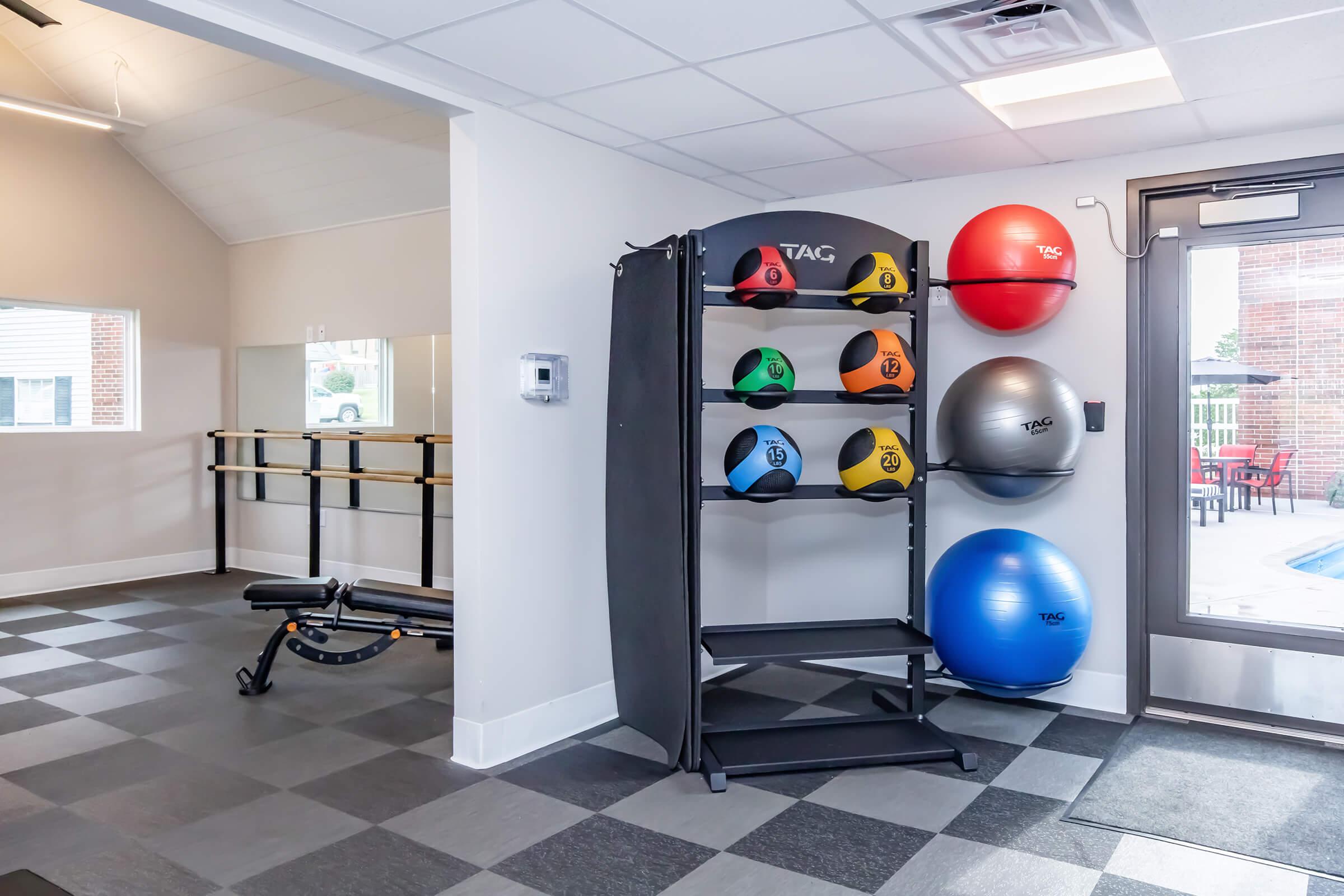 A fitness area featuring a rack of colorful exercise balls, a bench press, and a mirrored wall. The space is well-lit with large windows and has a modern design, emphasizing a healthy, active lifestyle.