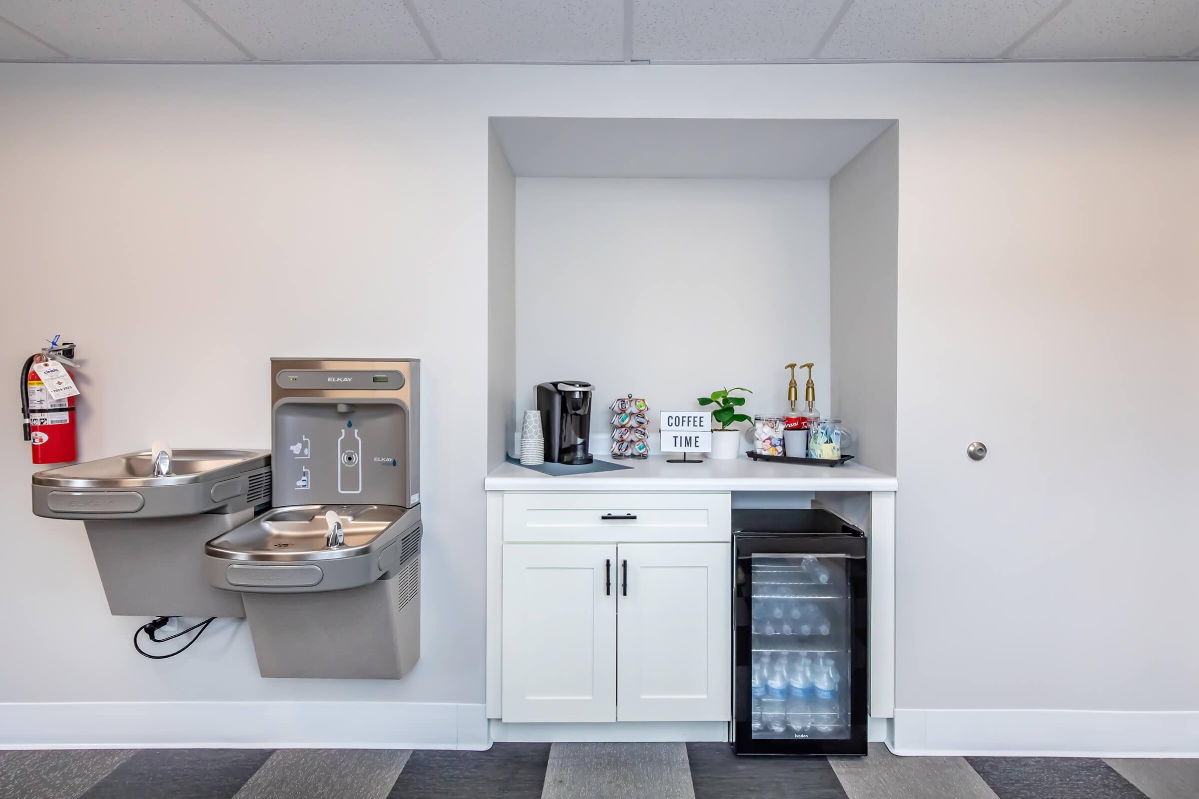 A clean break room area featuring two drinking fountains, a coffee machine, utensils, and a small refrigerator stocked with water bottles. A sign reads "Coffee Time," emphasizing the space's purpose for refreshments. The walls are painted in neutral colors, with modern flooring.