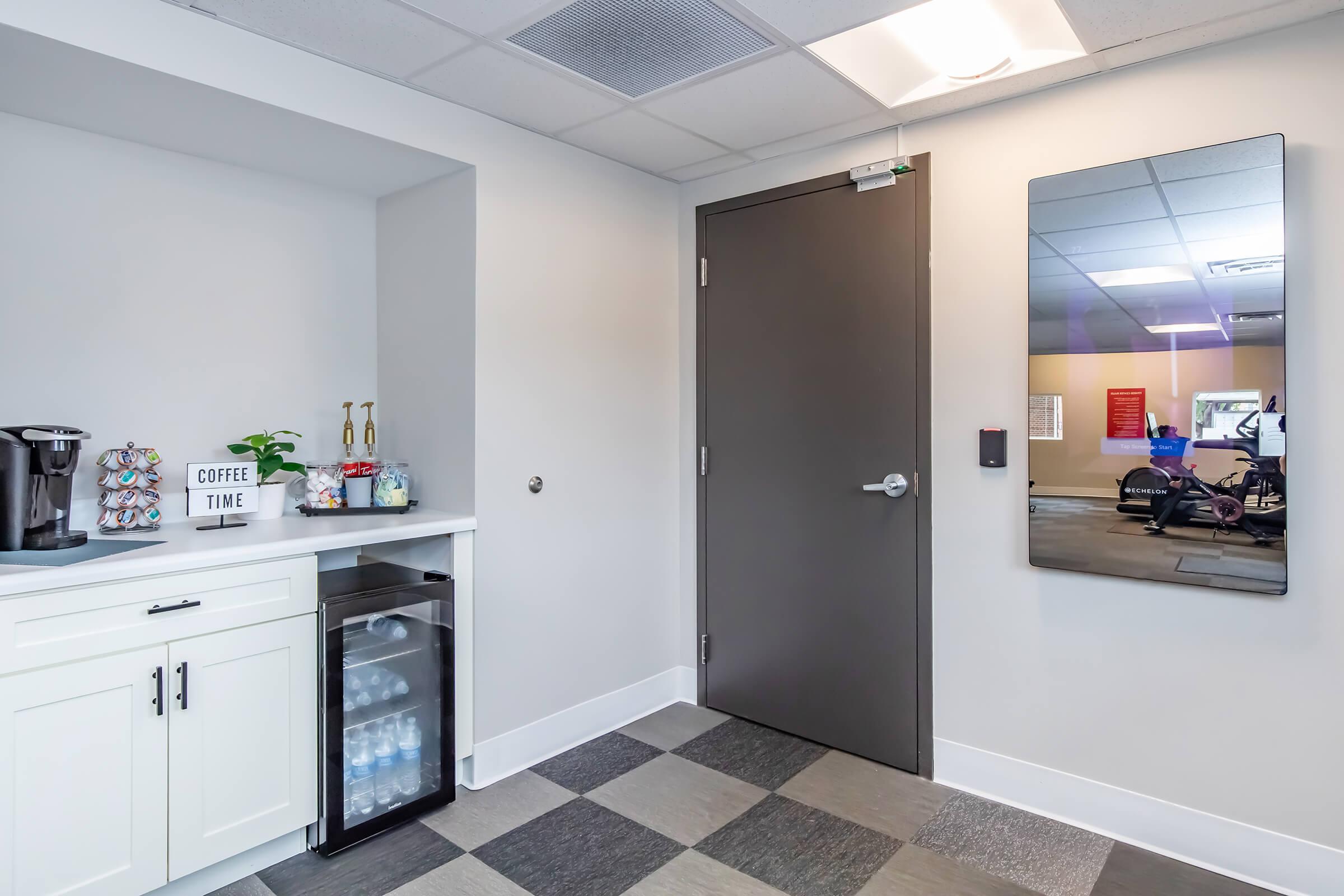 A modern break room featuring a coffee station with a coffee maker and decorative items, a mini fridge stocked with bottled water, and a large mirror on the wall. A grey door leads to another space, with an exercise area visible through the glass in the background. The floor has a modern checkered design.