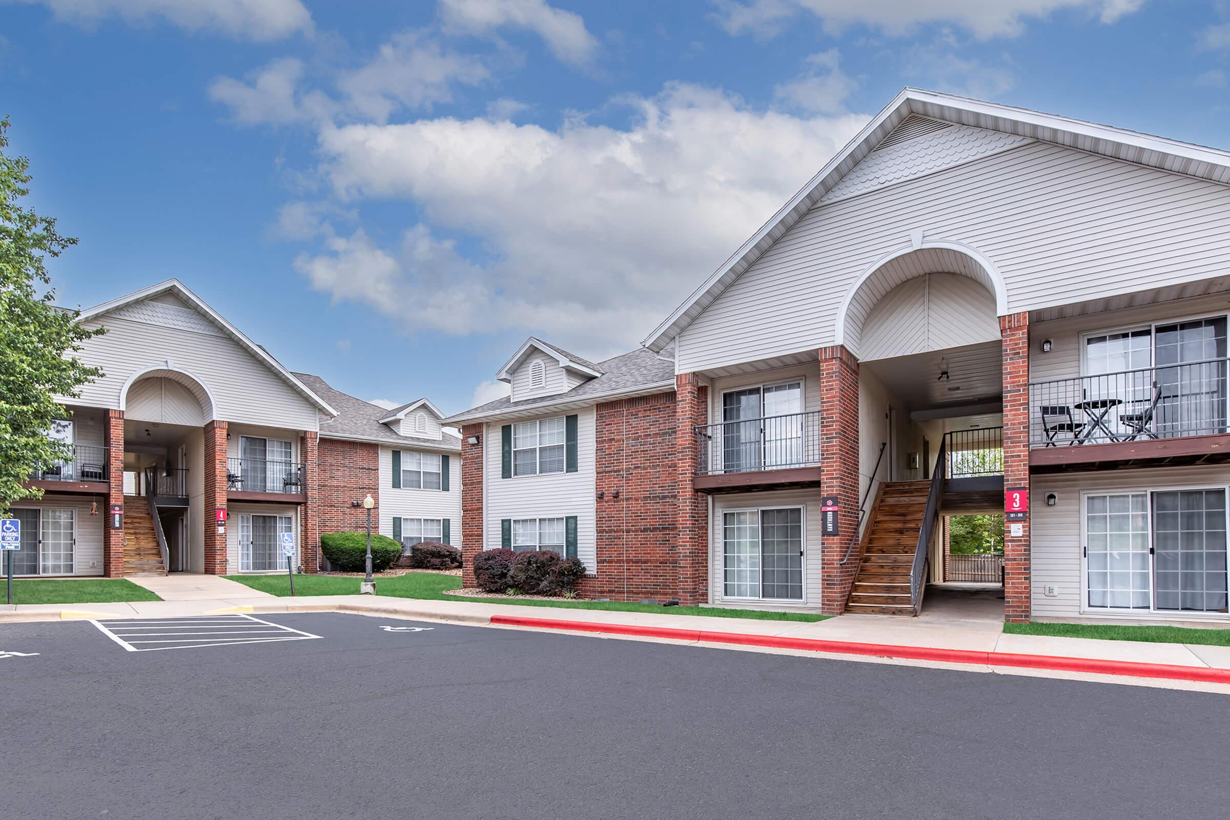 A view of a multi-story apartment complex featuring brick and siding exteriors. The buildings have balconies and stairways leading to the entrances. Lush green landscaping and a clear blue sky are visible in the background, creating a welcoming residential environment.