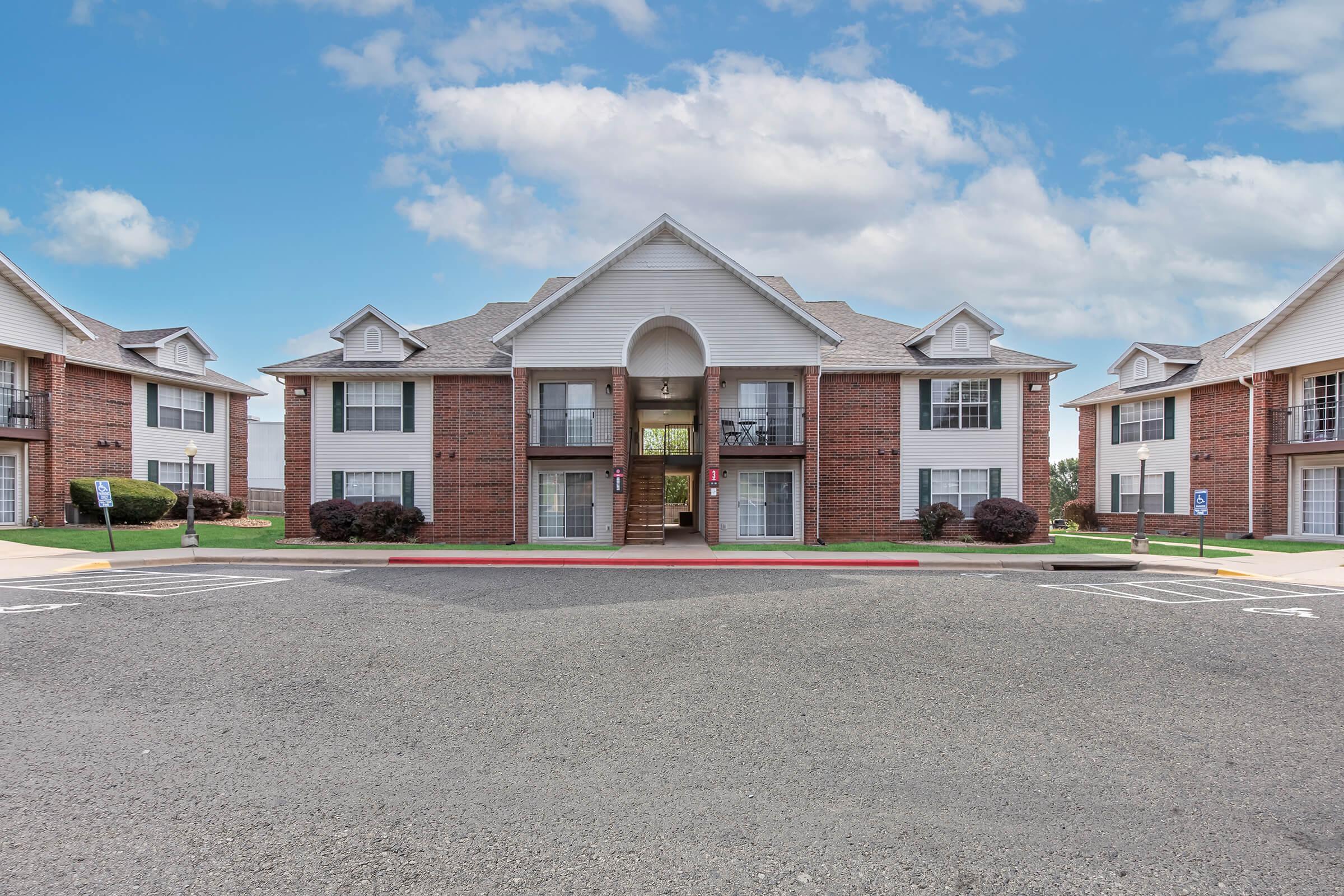 Two-story brick apartment building with white siding situated in a residential area. The entrance features a staircase, and there are several windows on the upper and lower levels. Surrounding the building are green lawns and parking spaces, all under a clear blue sky with a few clouds.
