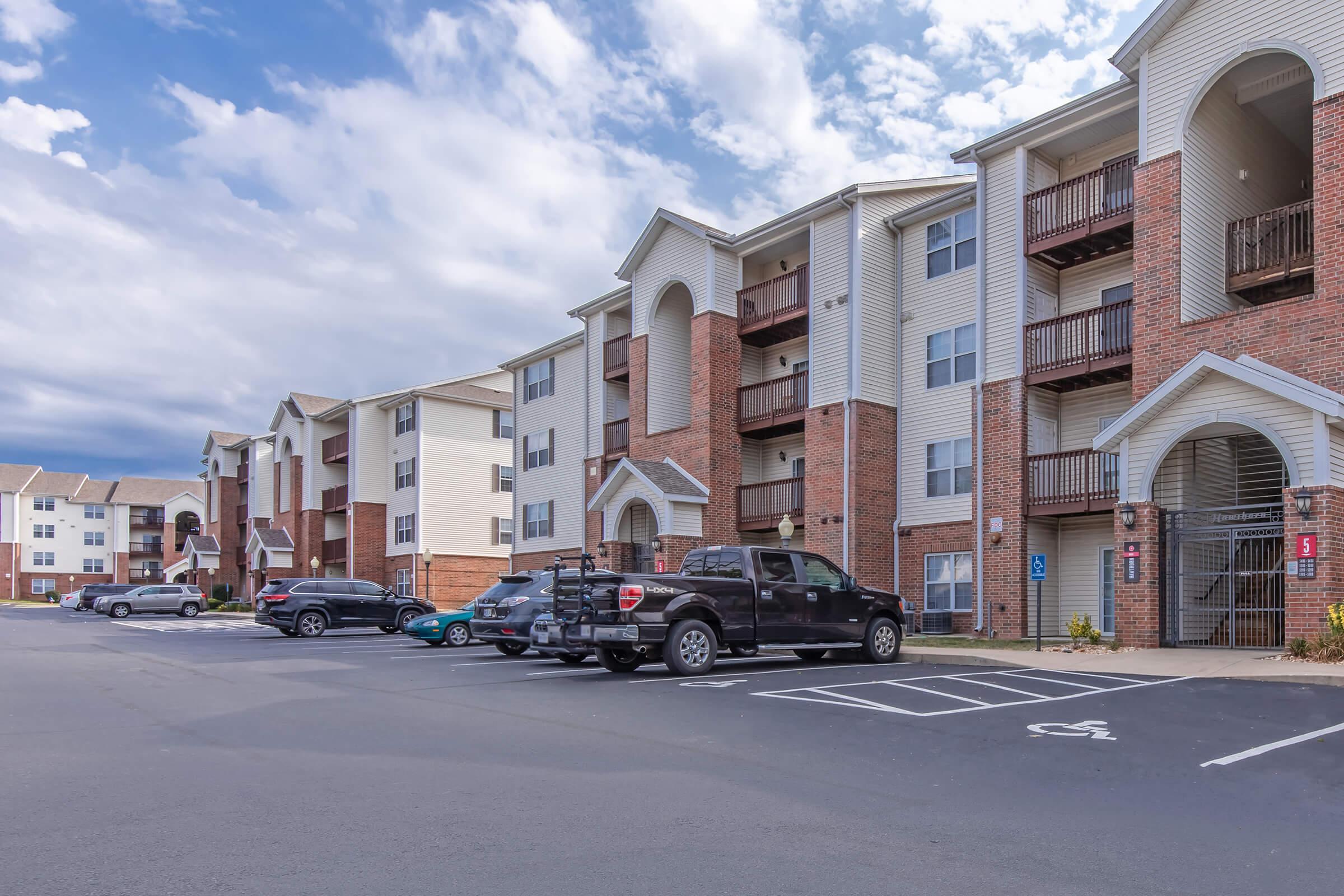 A parking lot lined with several apartment buildings. The foreground features a black pickup truck parked in a designated spot, with additional cars in the background. The sky is partly cloudy, creating a bright atmosphere for the residential setting.