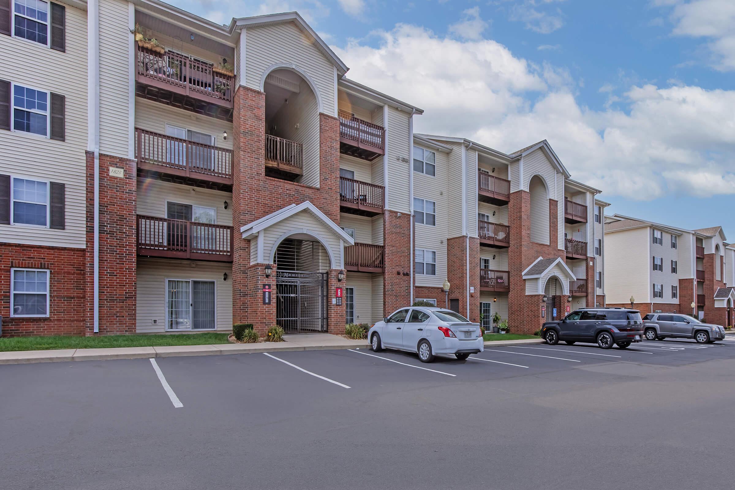View of a residential apartment complex featuring multiple buildings with balconies. The foreground includes a parking lot with several parked cars and well-maintained landscaping. The sky is partly cloudy, suggesting a pleasant day.