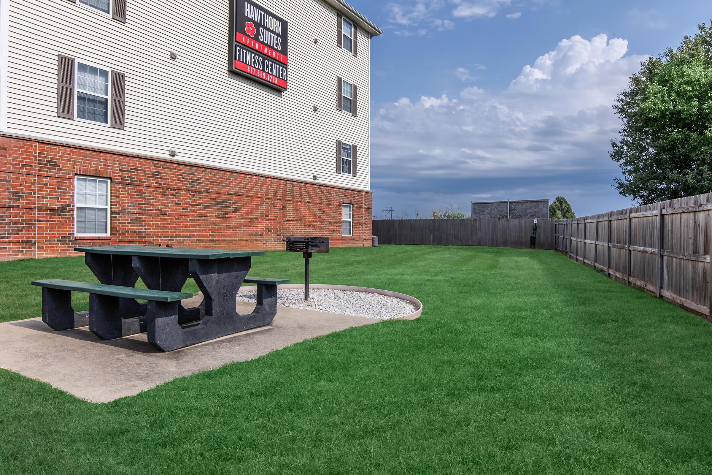 A grassy area featuring a picnic table on a concrete slab, surrounded by a gravel border. In the background, a wooden fence encloses the space, and an apartment building with a sign for a fitness center is visible. The sky is partly cloudy, creating a calm atmosphere.