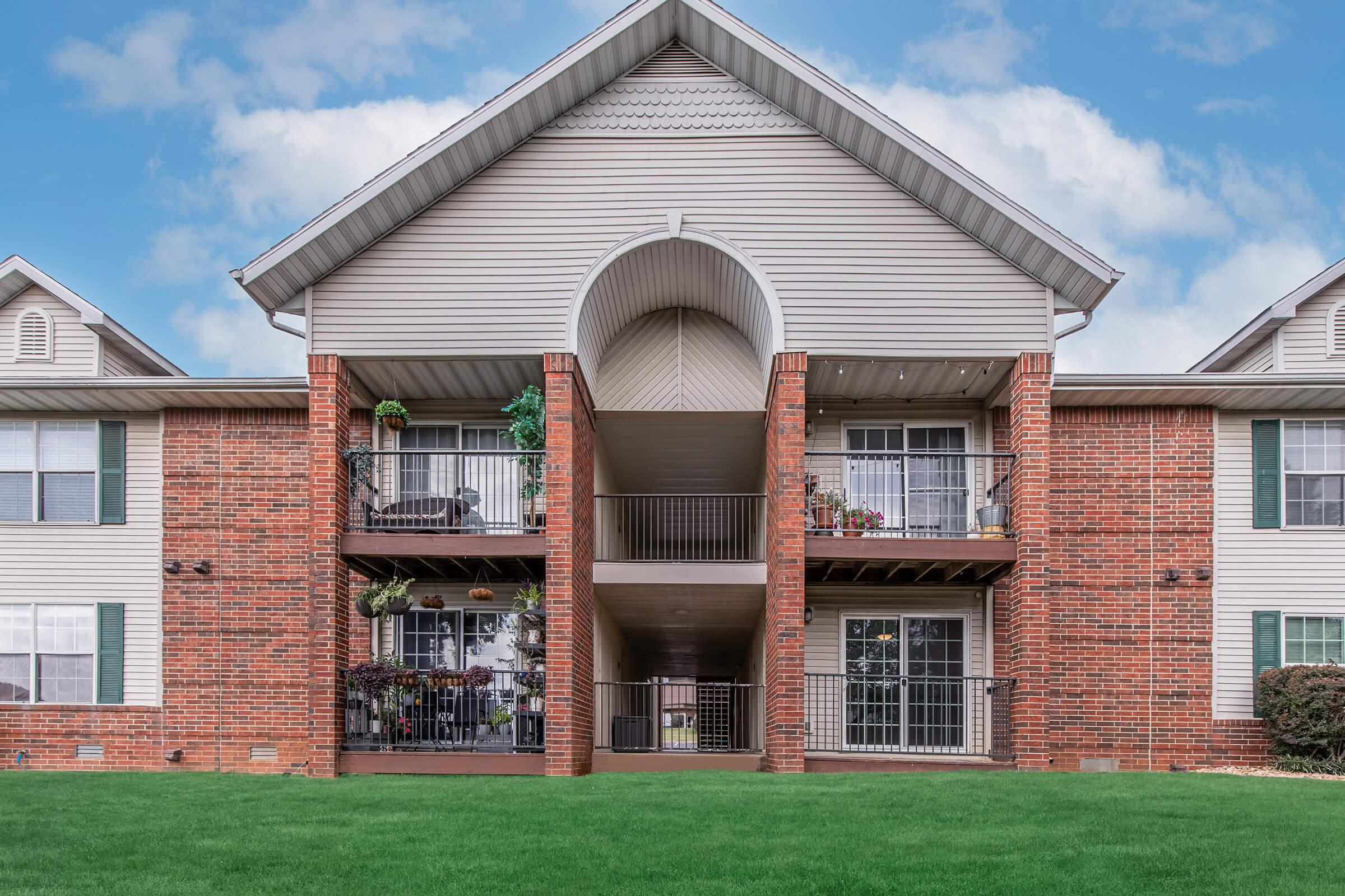 A multi-story apartment building with red brick and beige siding, featuring arched entryways and balconies. The building is surrounded by a lush green lawn and blue skies with fluffy clouds above.