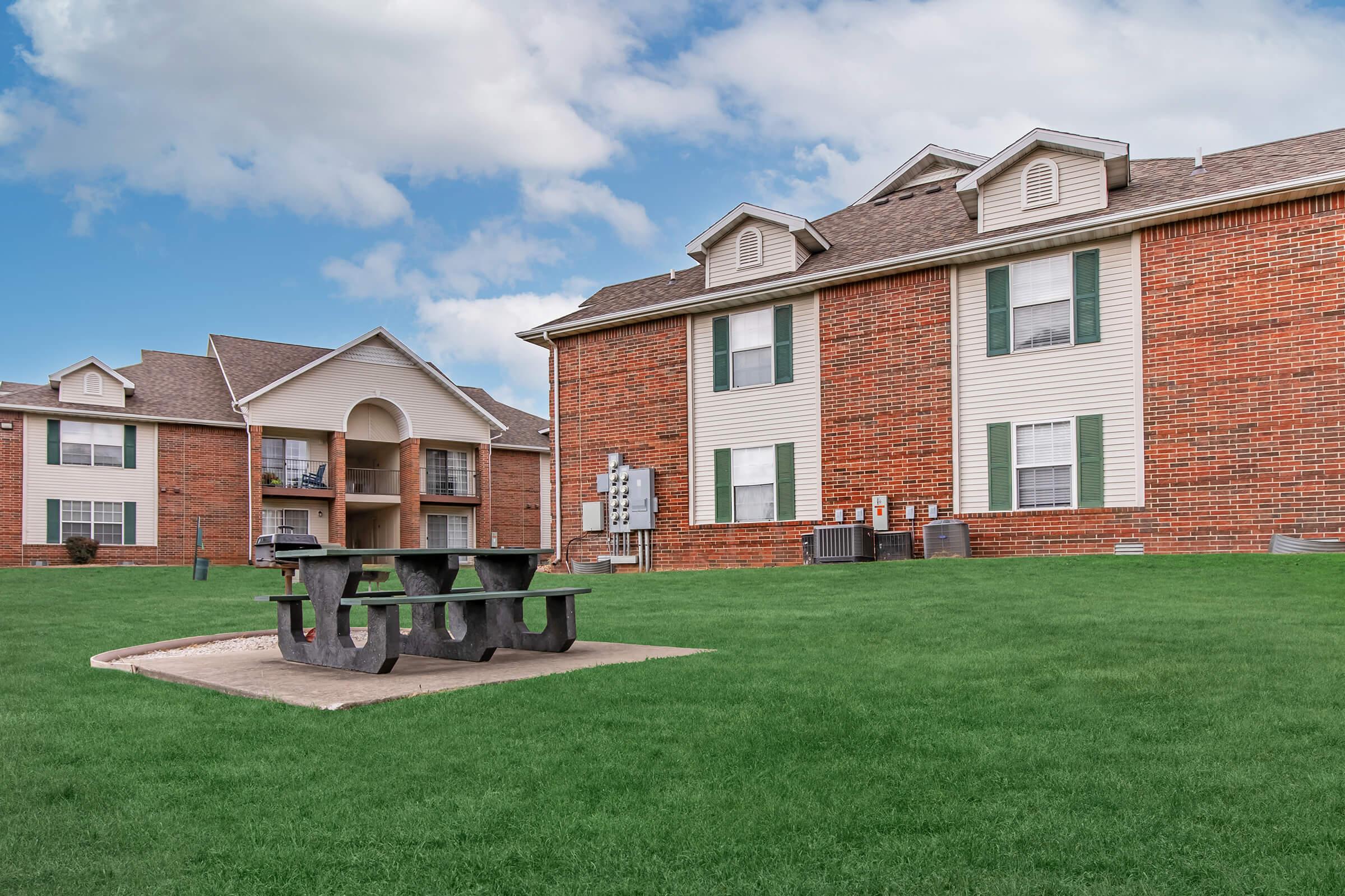 A view of a residential apartment complex featuring two buildings made of red brick and light-colored siding. The foreground shows a picnic table on a grassy area, with air conditioning units visible. The sky is partly cloudy, creating a bright, inviting atmosphere.
