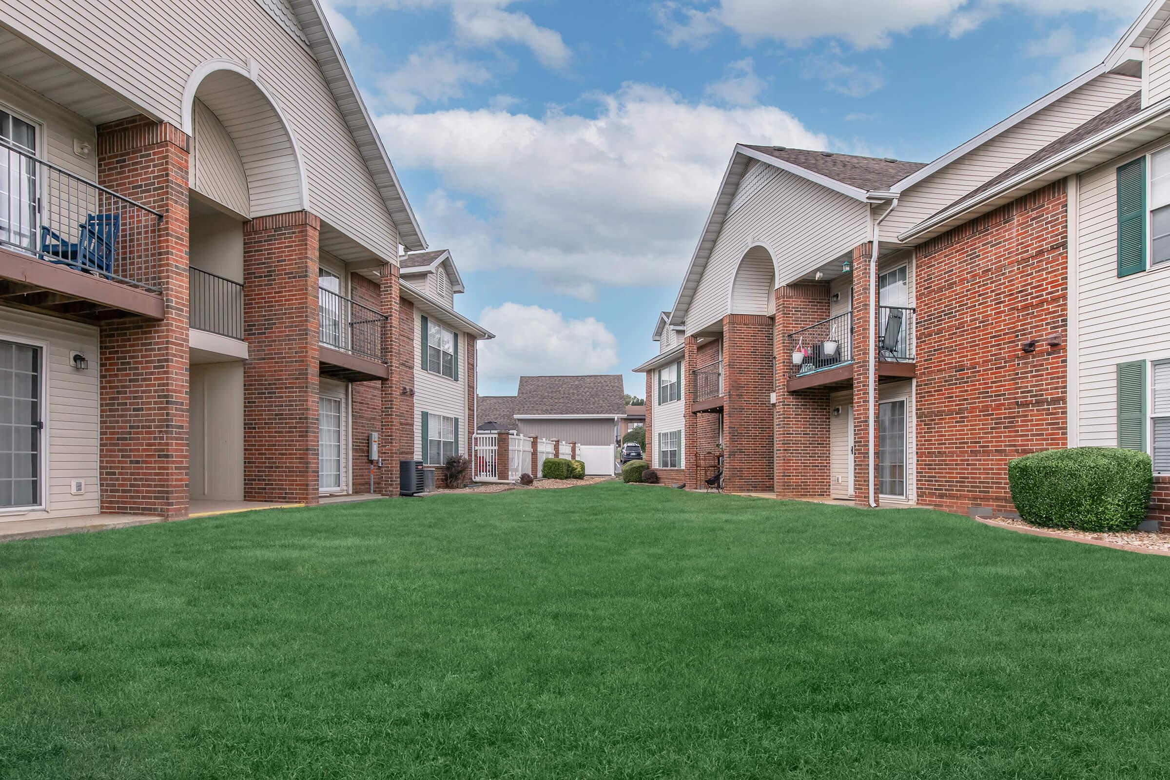View of an apartment complex courtyard featuring two buildings with brick facades on either side, surrounded by a well-maintained green lawn. The sky is partly cloudy, and there are balconies visible on the upper floors of the buildings.