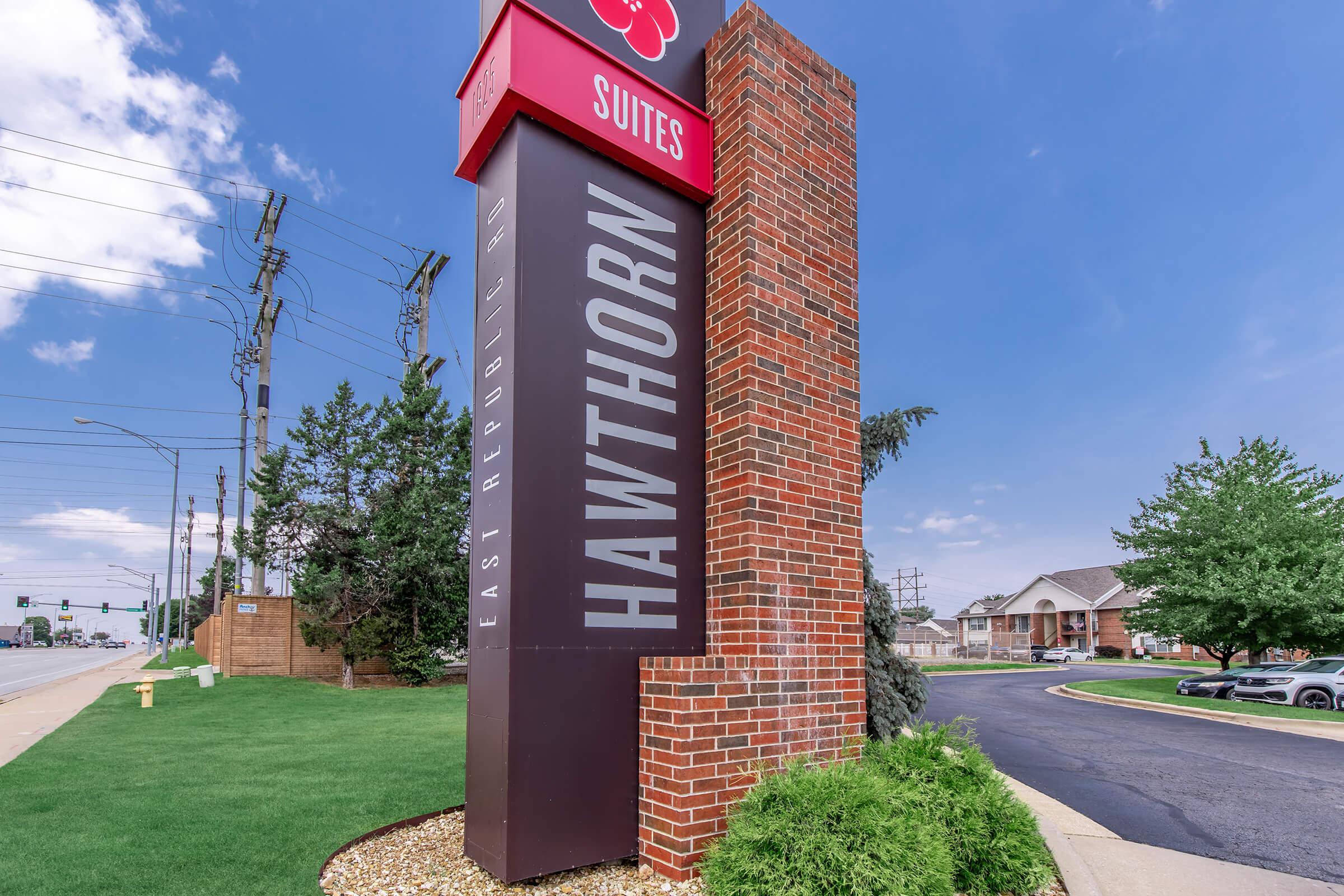 Sign for Hawthorn Suites located on East Republic Road, featuring red and black colors. The sign stands tall beside a landscaped area with grass and trees. Clear skies and a few clouds are visible in the background, indicating a sunny day. A road and vehicles are present nearby.
