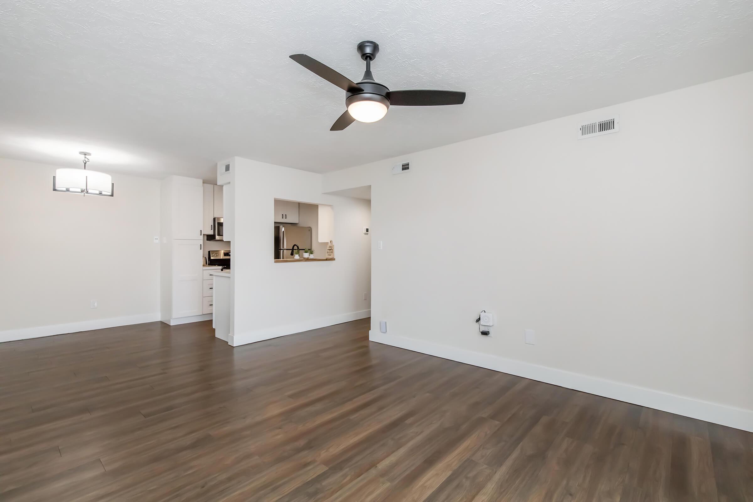 A spacious, well-lit living room featuring light hardwood flooring, a modern ceiling fan, and neutral-colored walls. In the background, a partially visible kitchen with white cabinetry and a dining area with a hanging light fixture. The space is open and airy, suggesting a contemporary design.