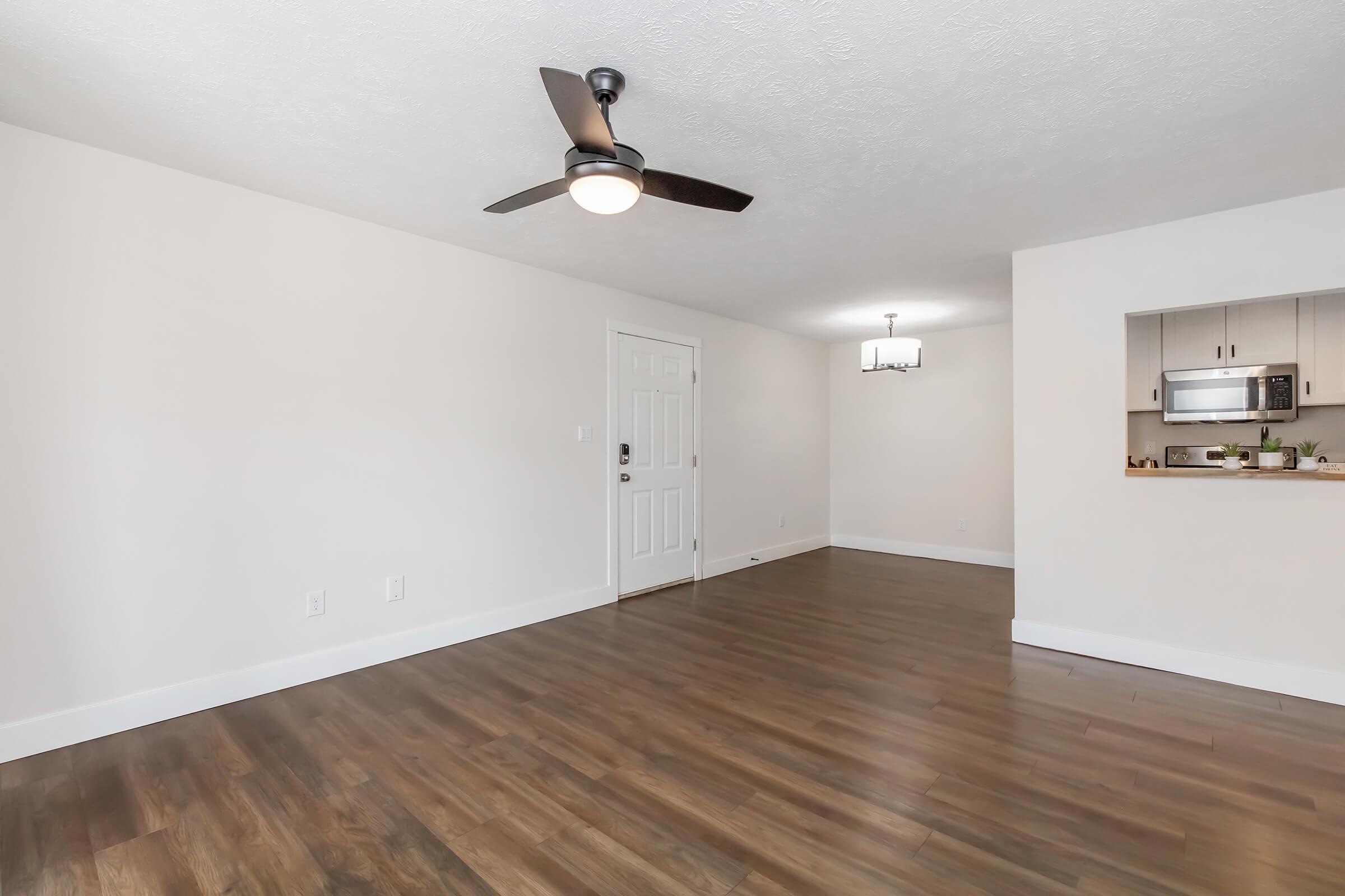 Spacious and bright living area featuring wooden flooring, a ceiling fan, and a neutral color palette. A doorway leads to another room, with an open kitchen area visible in the background, showcasing modern appliances and a small dining space. Overall, a contemporary and inviting atmosphere.
