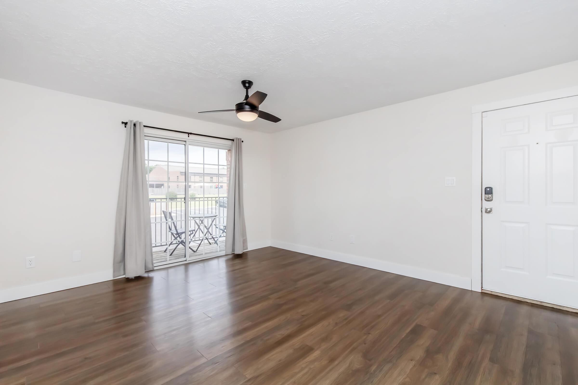 Interior view of a spacious living room with light wood flooring, a ceiling fan, and a large sliding glass door leading to a balcony. The walls are painted in a neutral color, and there are light curtains at the window. A front door is visible on the right side of the image.