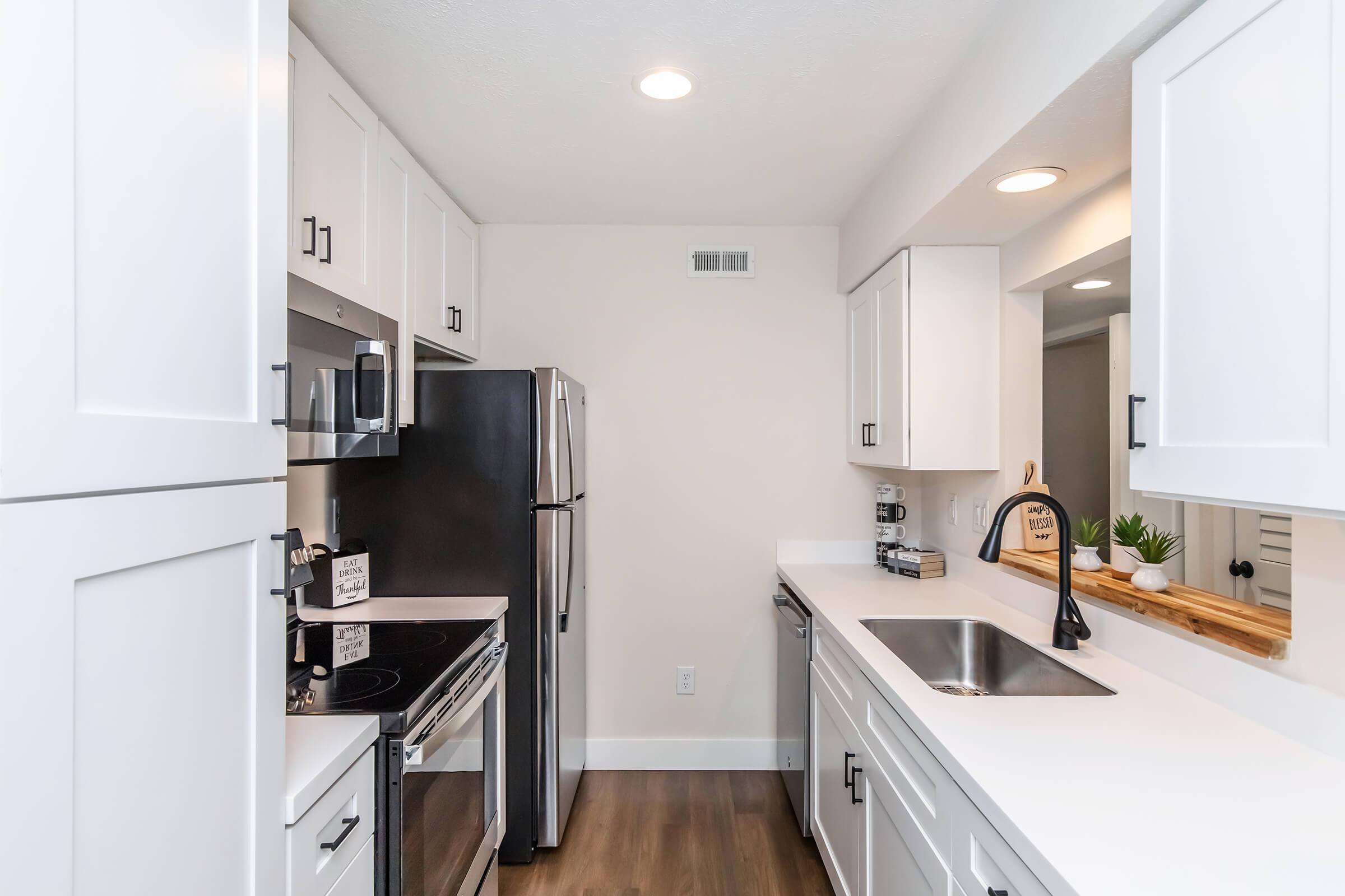 Modern kitchen featuring white cabinetry, stainless steel appliances, and a sleek countertop. Includes a sink, oven, microwave, and coffee maker. The space is bright with recessed lighting and a minimalist design, complemented by a wooden shelf displaying small plants and decor.