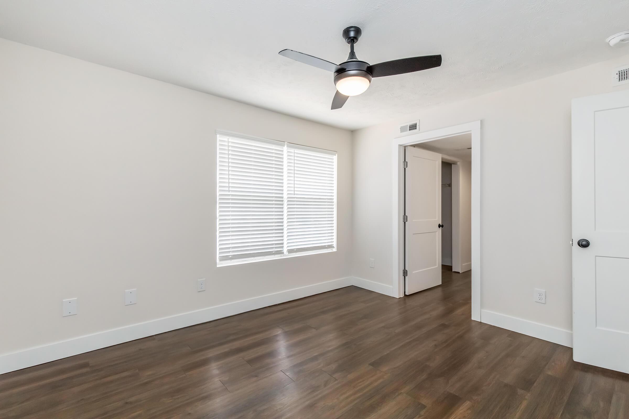 A clean and minimalistic room featuring light beige walls, a ceiling fan, and dark hardwood flooring. A window with white blinds allows natural light, and a door leads to another room. The space is unoccupied, showcasing a modern design with a bright and airy feel.