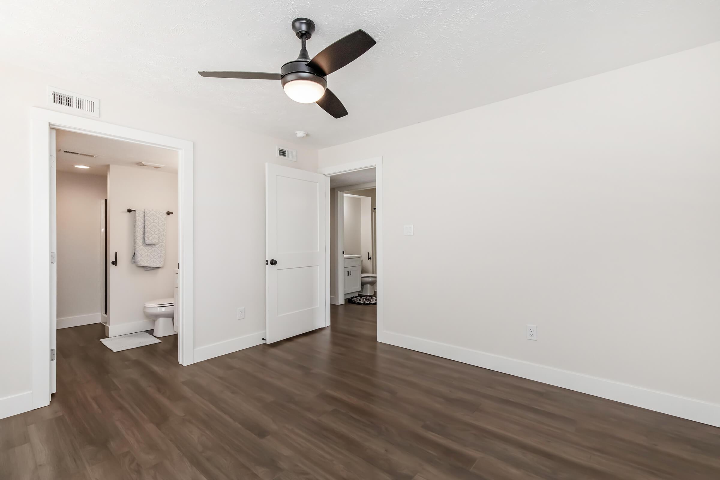A spacious, well-lit room with light-colored walls and brown wooden flooring. A ceiling fan is visible, and there are two doorways leading to other areas, one of which has a bathroom visible in the background. The overall decor is simple and modern.