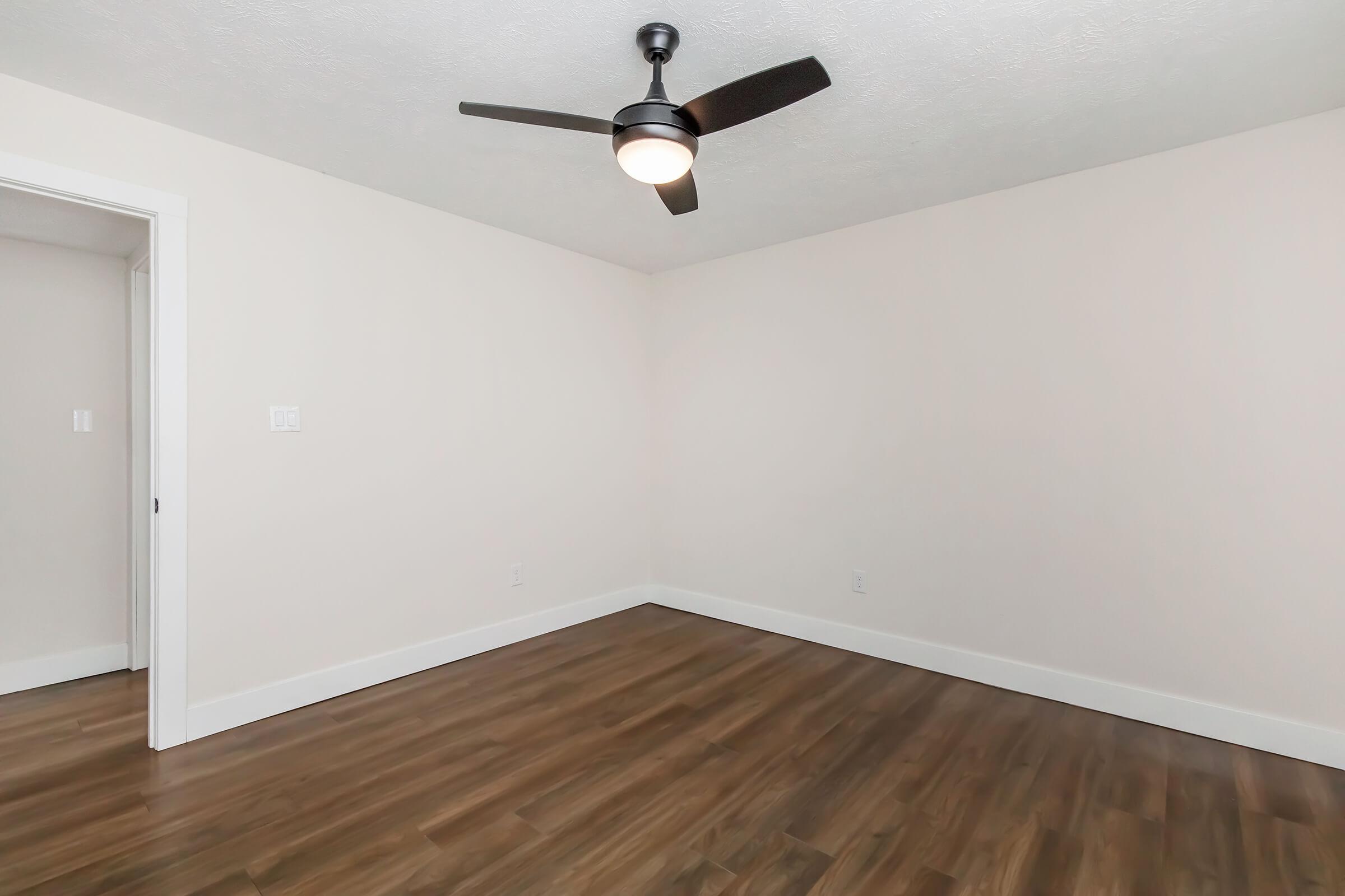 A spacious, empty room with light-colored walls and a modern ceiling fan. The flooring is wooden, and there is a doorway visible on the left side. The room is well-lit and has a clean, minimalist design, making it suitable for various uses.