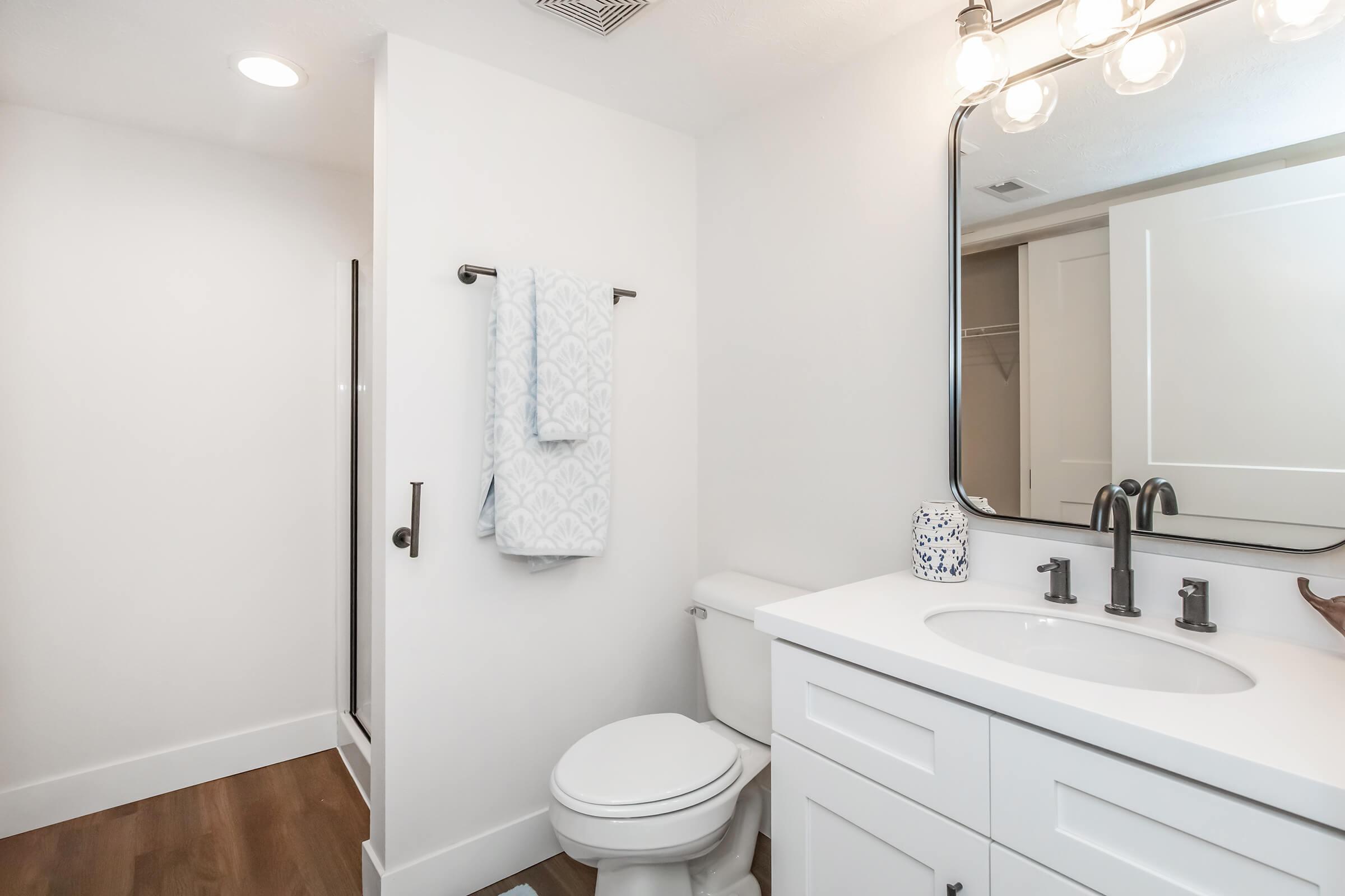 A modern bathroom featuring a white vanity with a sink, a large mirror, and stylish fixtures. There is a toilet, a towel rack with a light blue towel, and a walk-in shower. The walls are painted white, and the flooring is wooden, creating a clean and bright atmosphere.