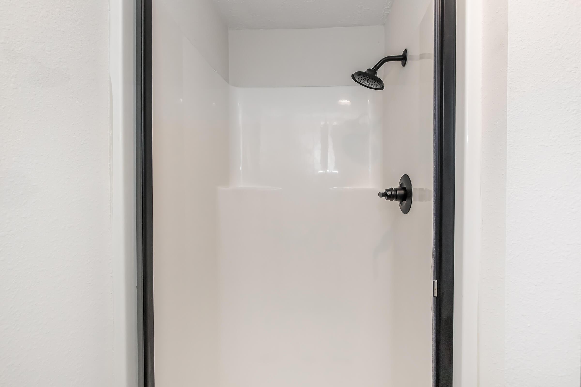 A clean, modern shower with a black showerhead and a smooth white interior. The shower space includes a black handle and is enclosed by a black frame, set against a plain white wall. The overall design is minimalist and contemporary.