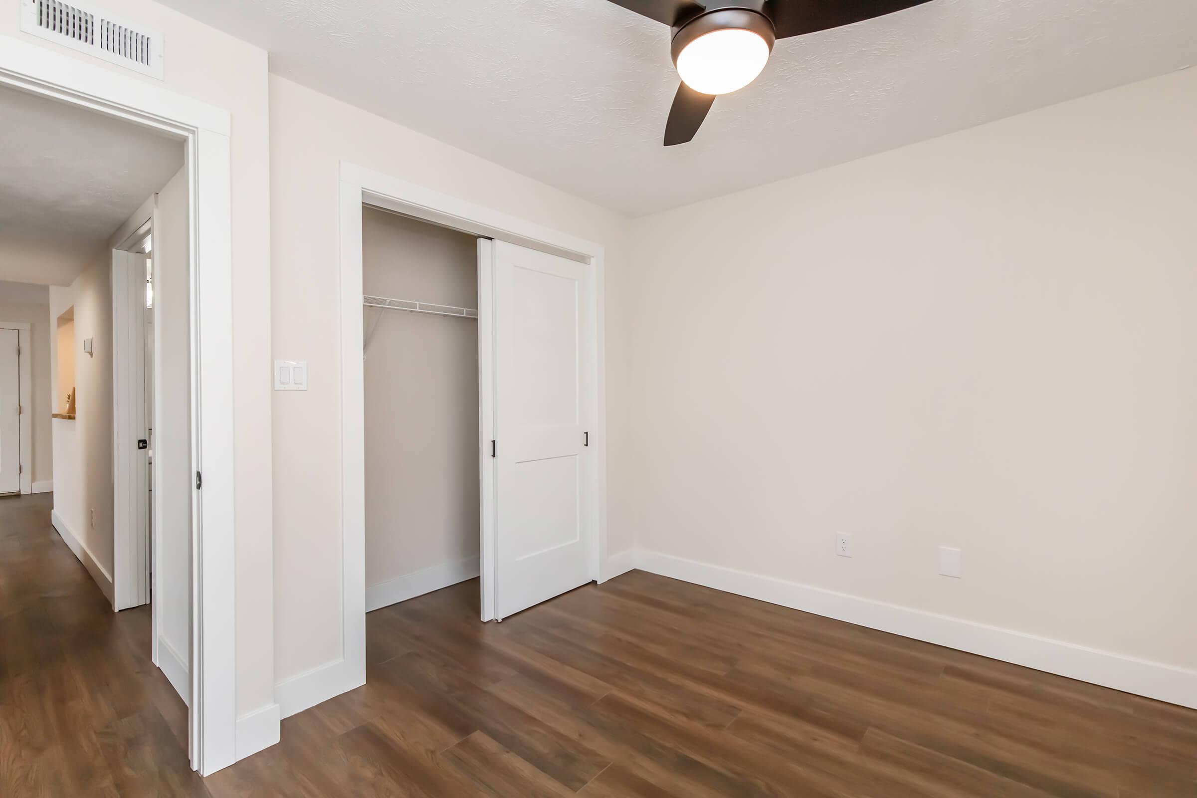 A well-lit interior view of a room featuring light-colored walls and a ceiling fan. The room has a closet with double doors slightly ajar, and a hardwood floor reflecting natural light. Visible in the background is a hallway leading to other areas of the home.