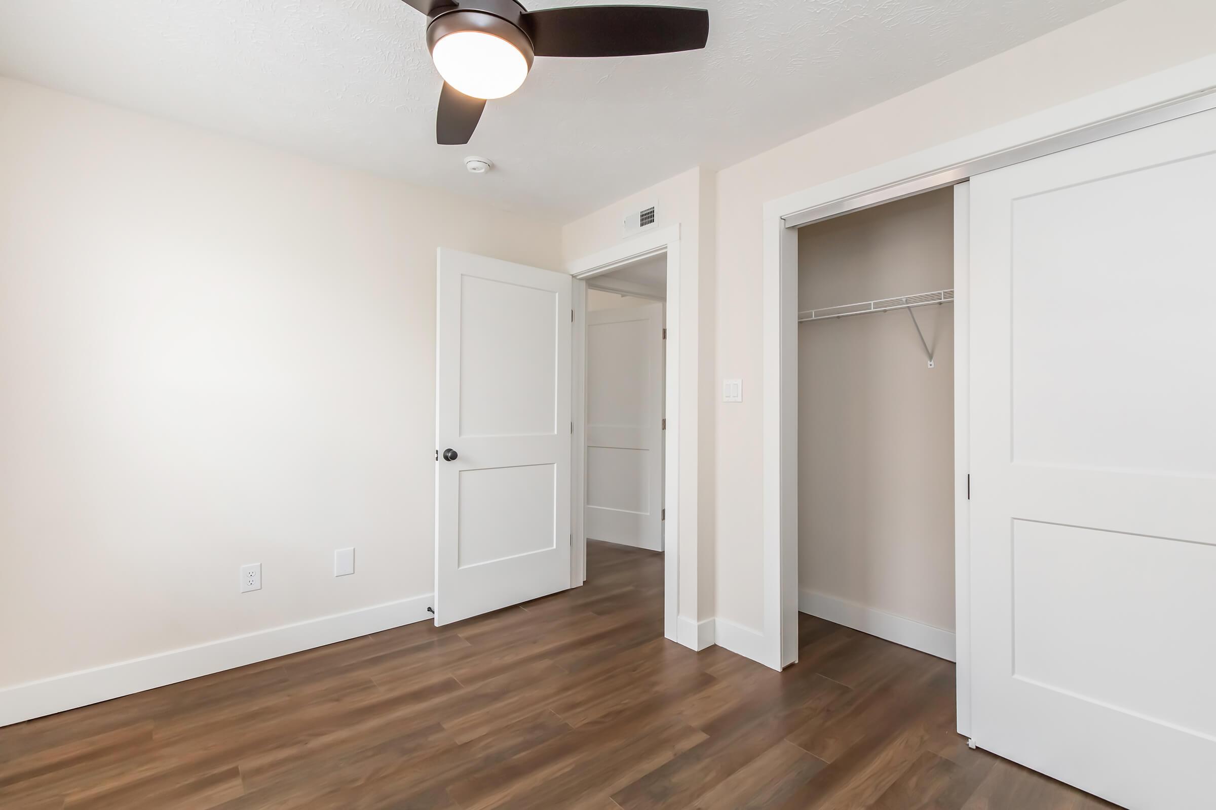 A bright, empty room featuring light-colored walls, a ceiling fan, and hardwood flooring. One door leads to a closet, while another door is partially open, suggesting access to another area. The space is well-lit with natural light coming from an unseen window.