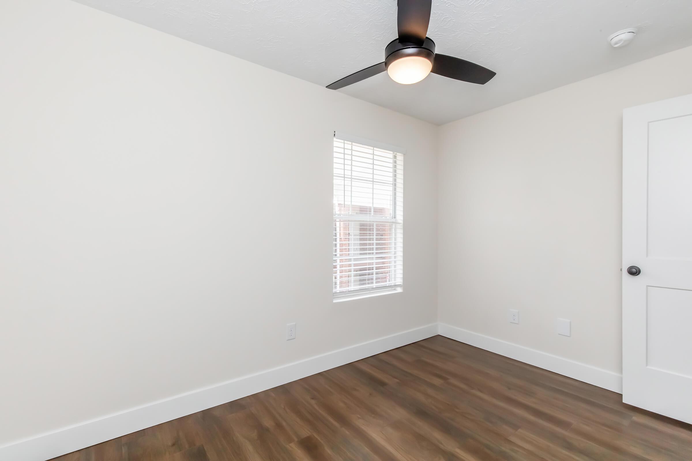 A bright, empty room with light-colored walls and a ceiling fan. It features a window with blinds allowing natural light in and a hardwood floor. The room has a simple, modern aesthetic, suitable for various uses, such as a bedroom or office.